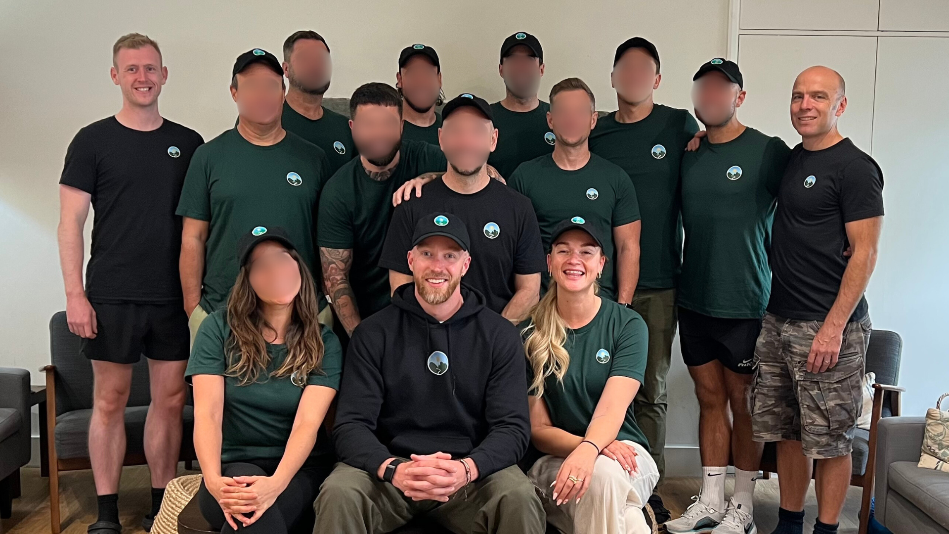 Group of 13 people, 11 men and 2 women, posing indoors with some wearing matching green and black shirts and black caps, smiling for a photo.