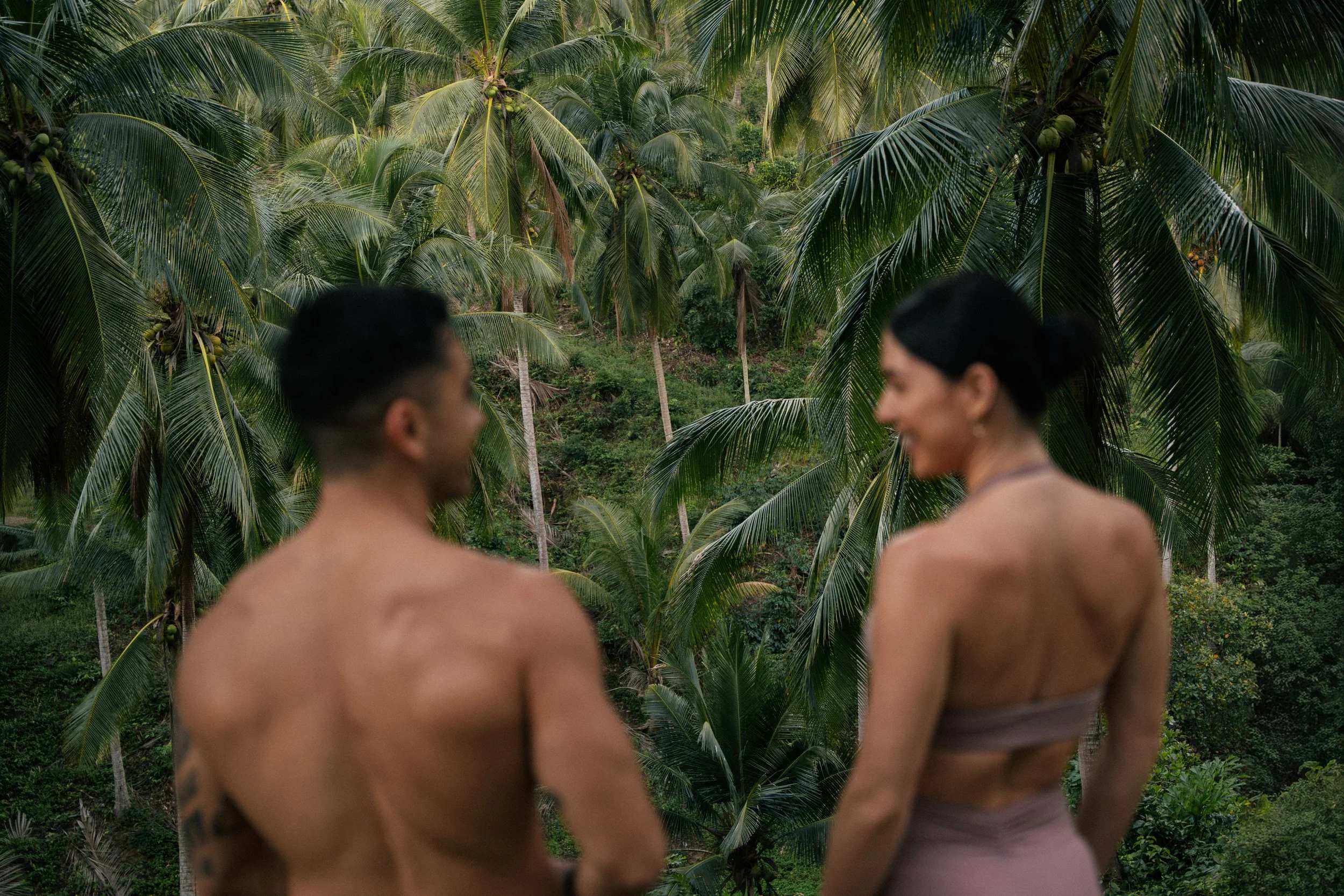A man and woman standing shirtless, turned towards each other and smiling, in front of a lush tropical background with numerous palm trees.