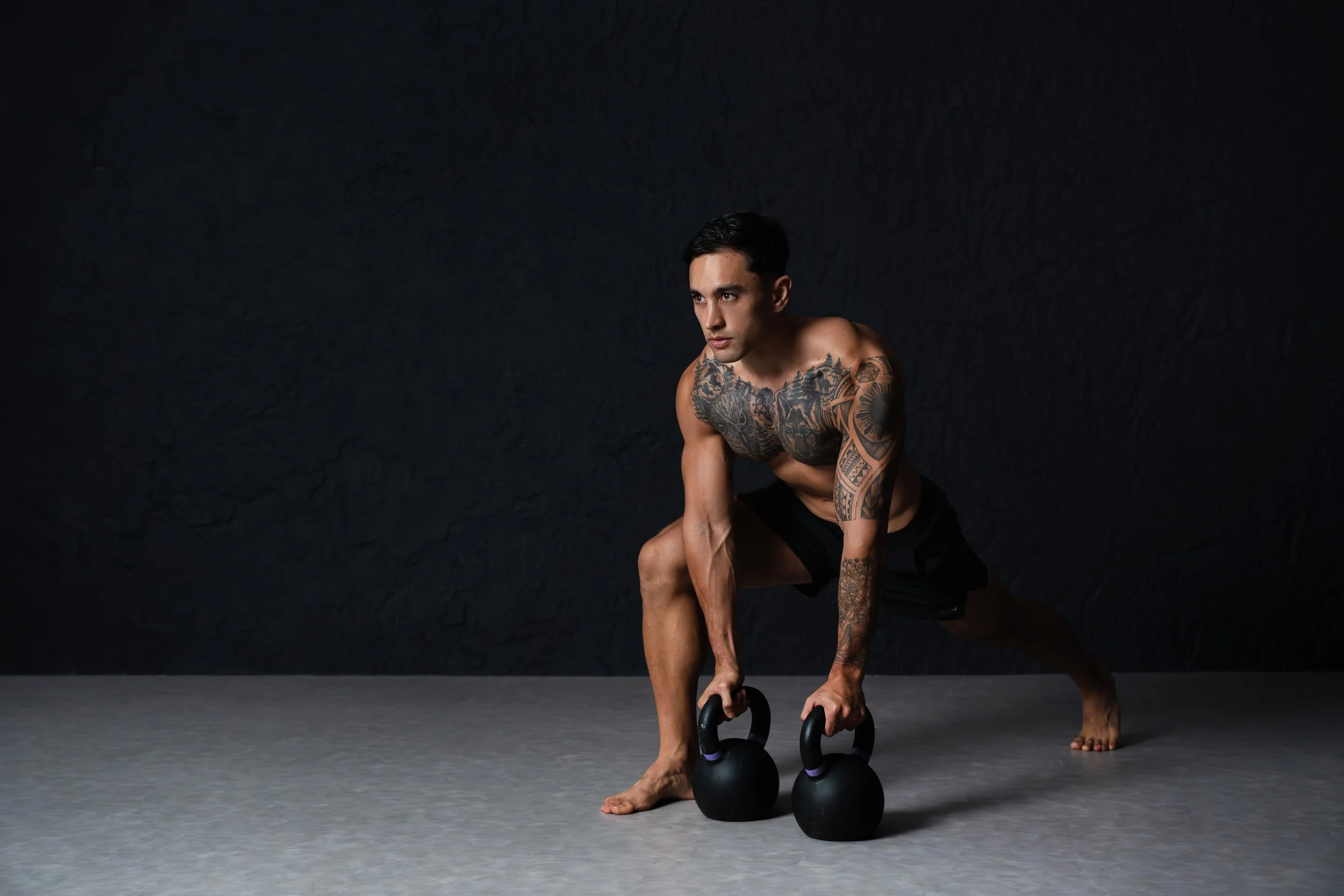 A shirtless man with tattoos on his chest and arms performing a workout with two kettlebells on a gray floor against a black background.