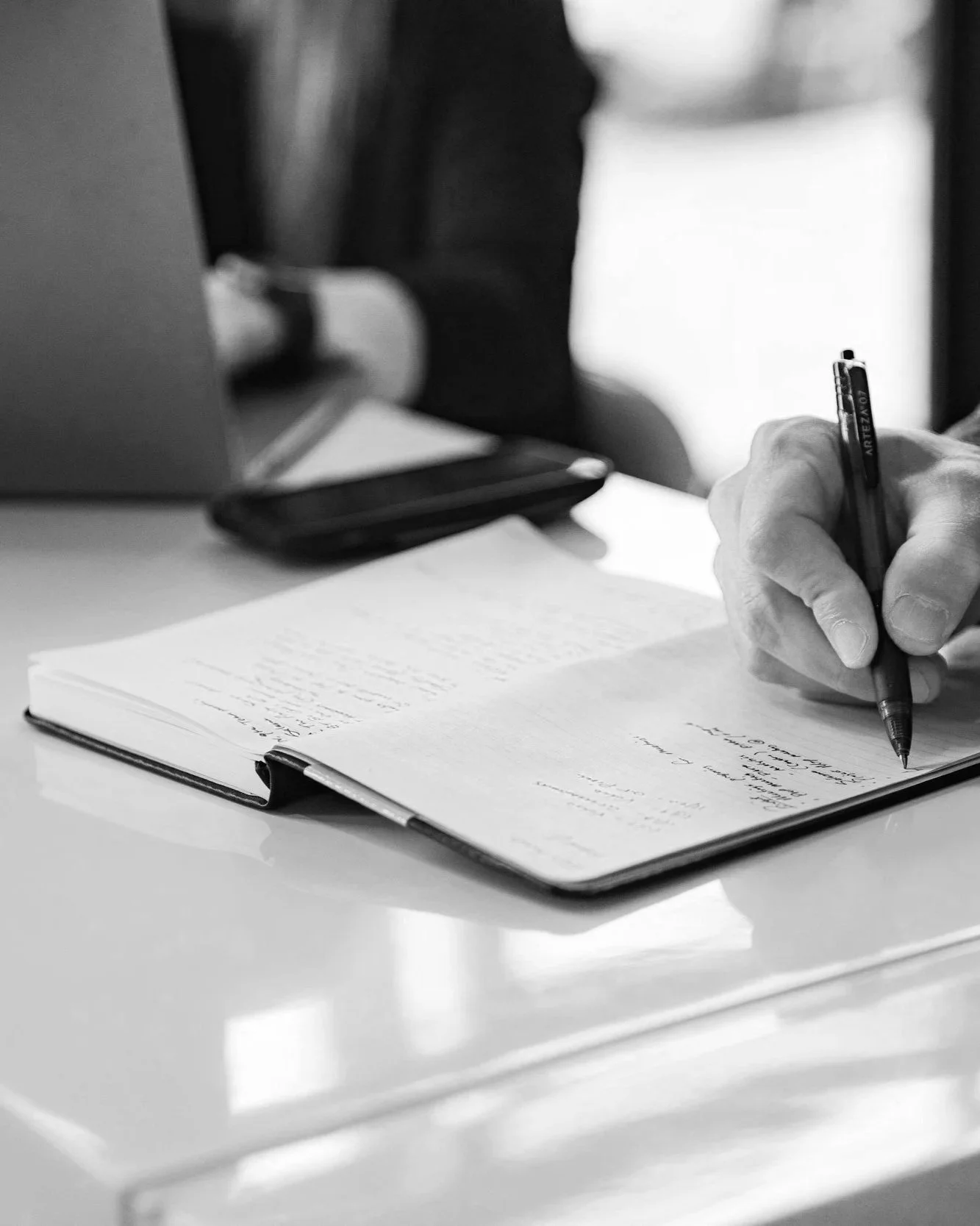 A person writes in a notebook at a desk with a pen, a laptop, and a mobile phone nearby in a black and white photo.