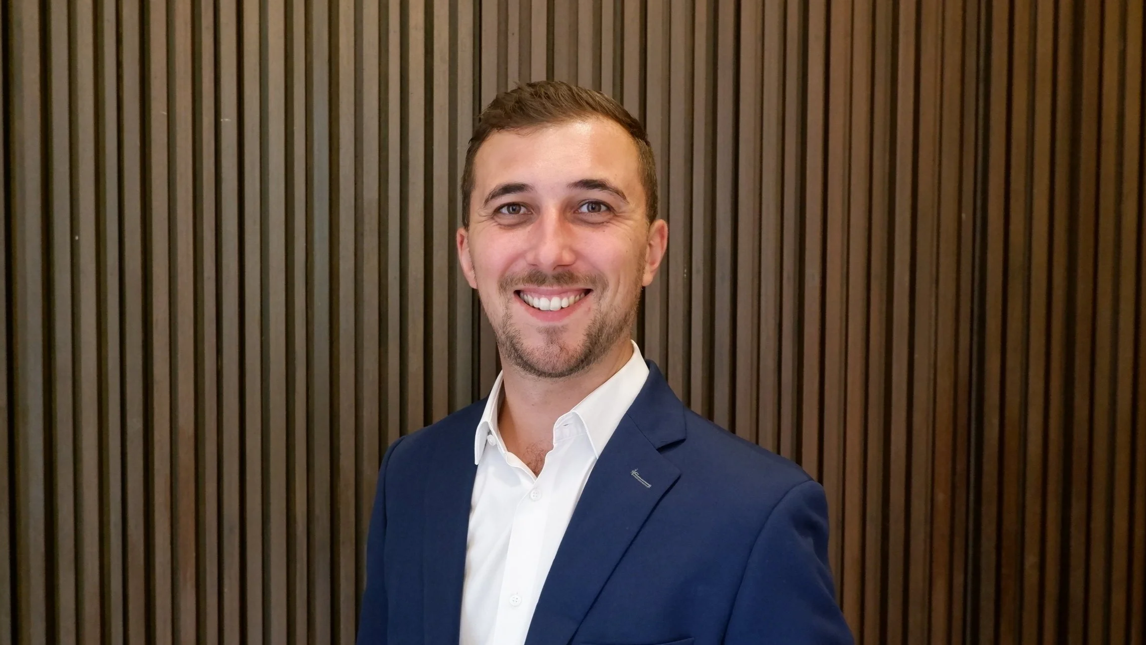 A man in a blue suit and white shirt smiling against a wooden vertical slat background.