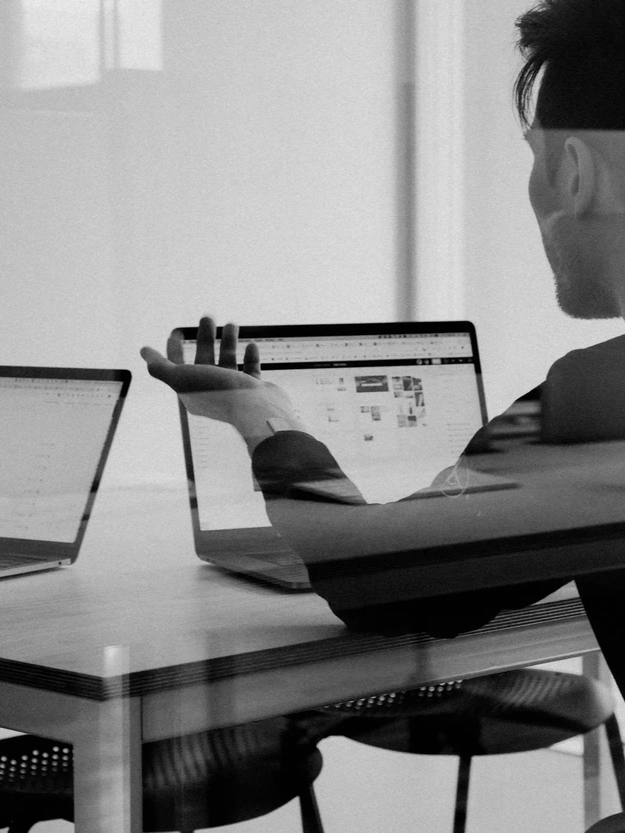 A person with glasses and a smartwatch sitting at a desk, looking at a laptop screen. The person is gesturing with their hand, and there is another laptop on the desk. The environment appears to be an office or meeting room.