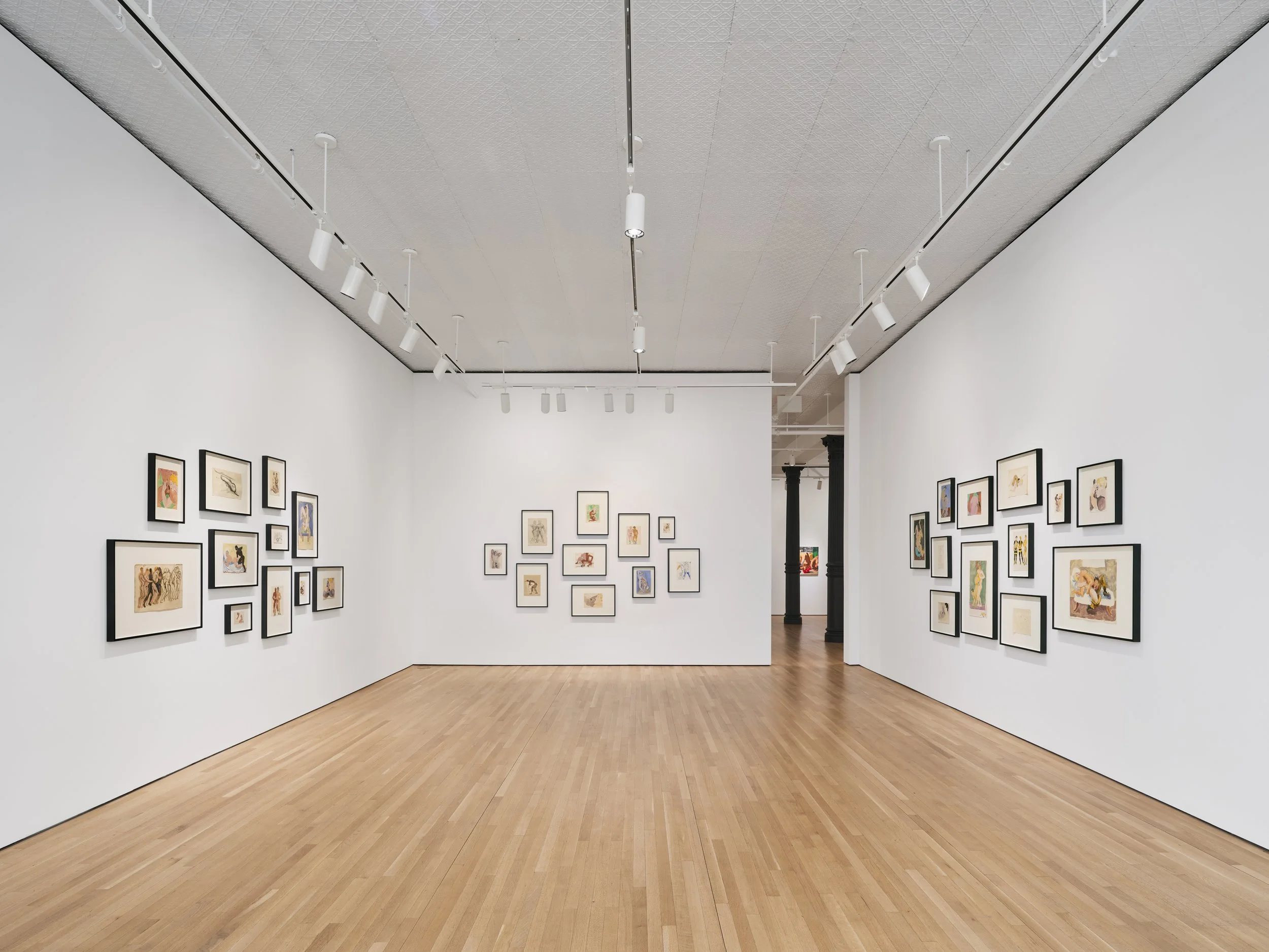 A modern art gallery with white walls and wooden floors, displaying multiple framed artworks arranged in clusters on the walls under ceiling track lighting.