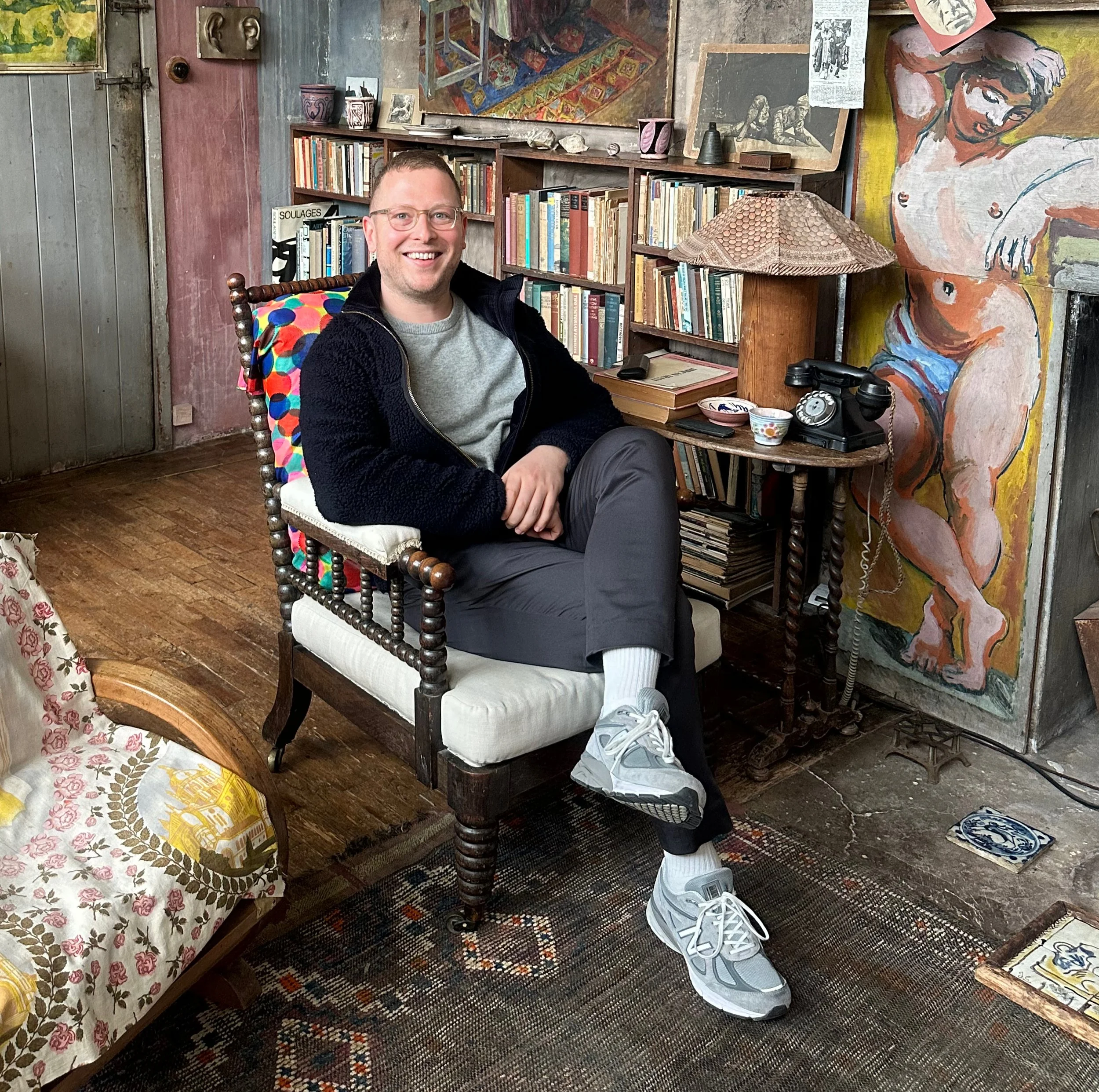 A man is sitting on a vintage wooden armchair in a cosy room filled with books and artwork. Smiling and wearing glasses, he is dressed in a grey jumper, dark trousers, pale trainers and a navy jacket. He has one leg crossed over the other.
