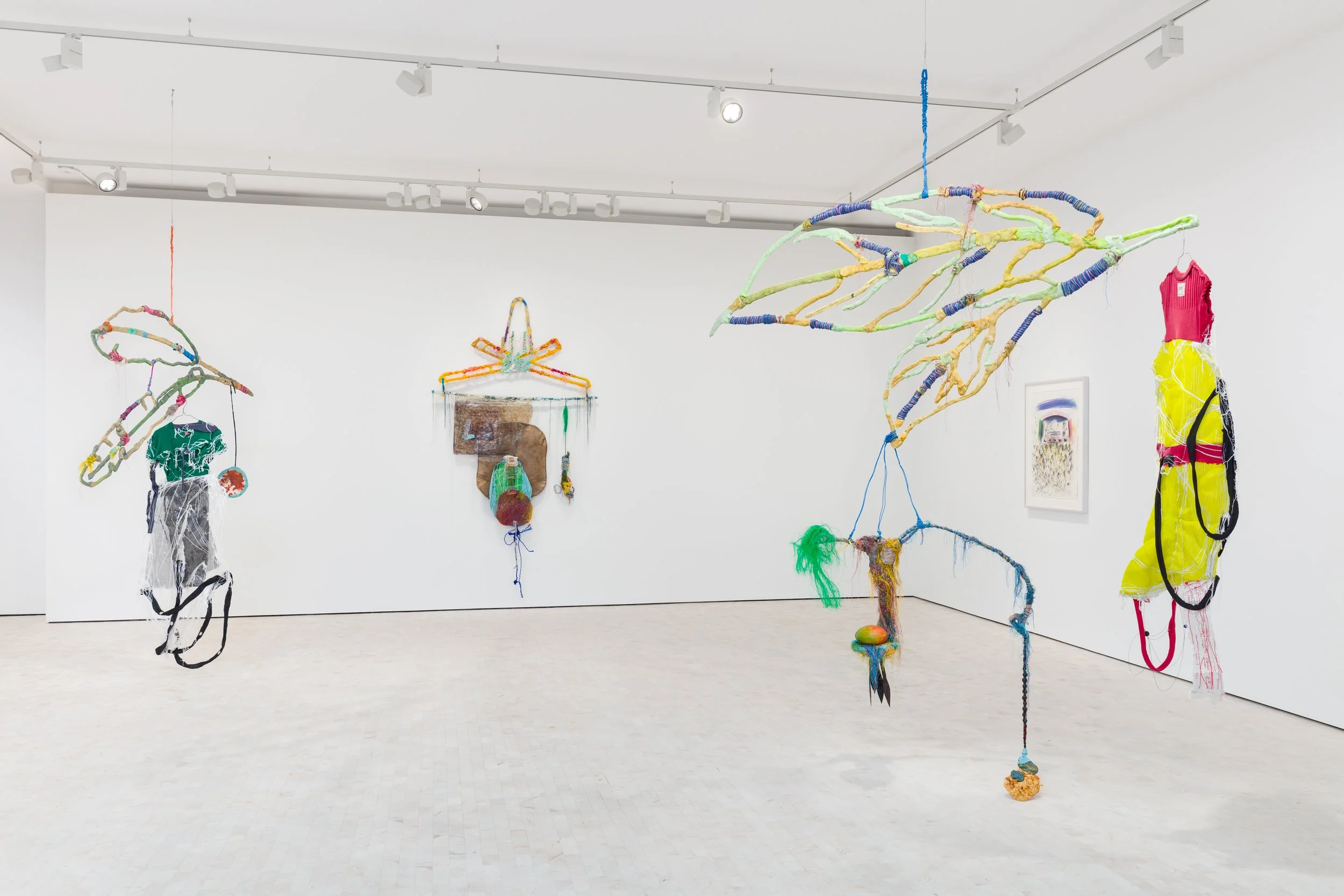 Colorful mixed-media art installation with hanging sculptures made of fabric, yarn, and other materials in a white gallery space.