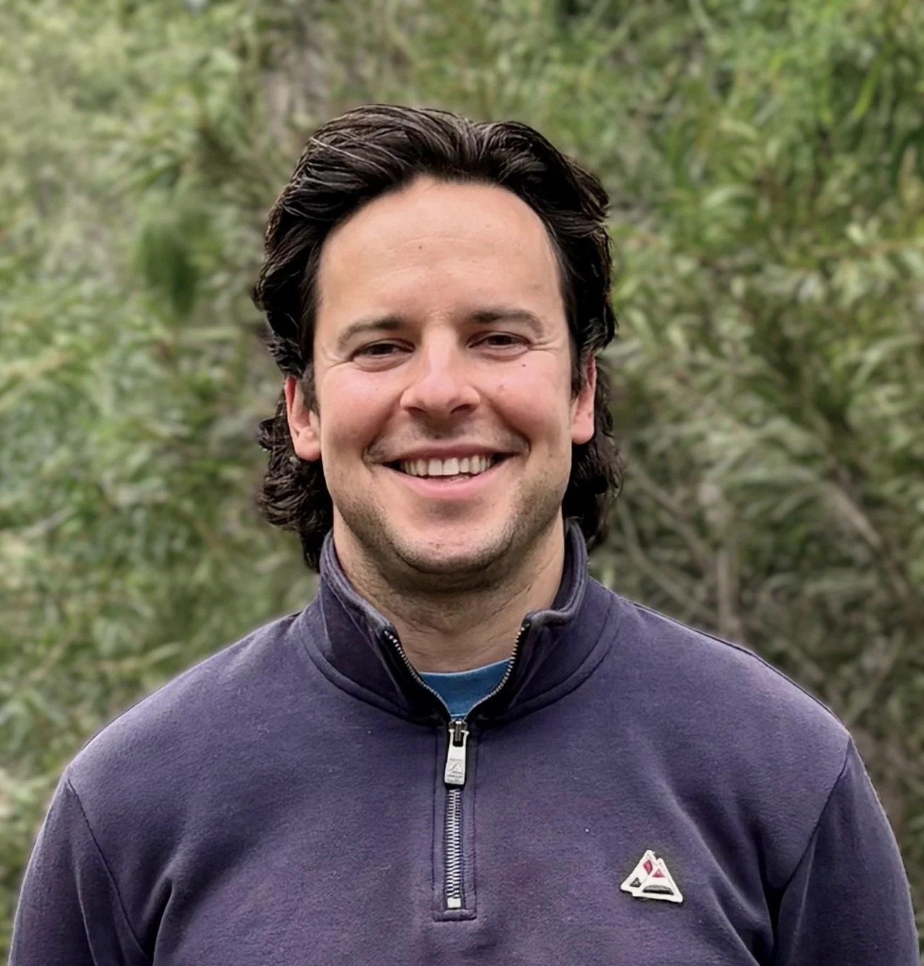 A man with dark hair smiling outdoors, wearing a dark zip-up jacket with a small logo on the chest, and a background of green foliage.