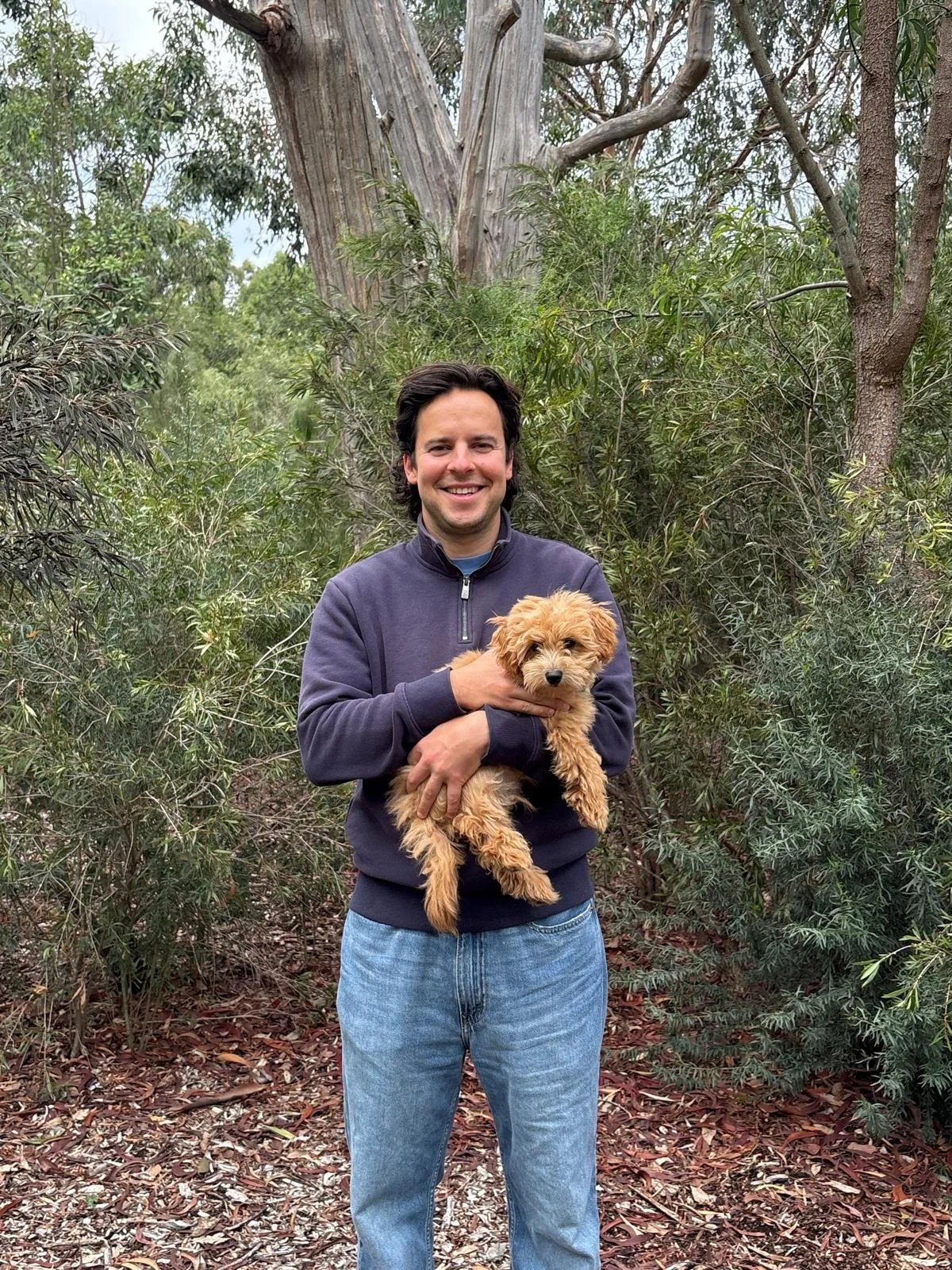 A man smiling in an outdoor forest setting, holding a small fluffy puppy.
