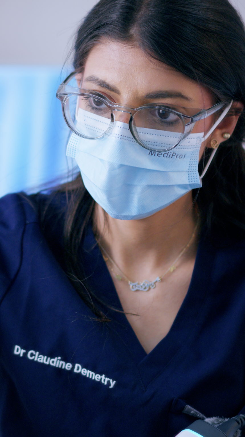 A woman with dark hair wearing glasses, a face mask labeled "MediproS," and a navy blue medical uniform. She has small earrings and a gold necklace, and her uniform has the name "Dr. Claudine Demetry."