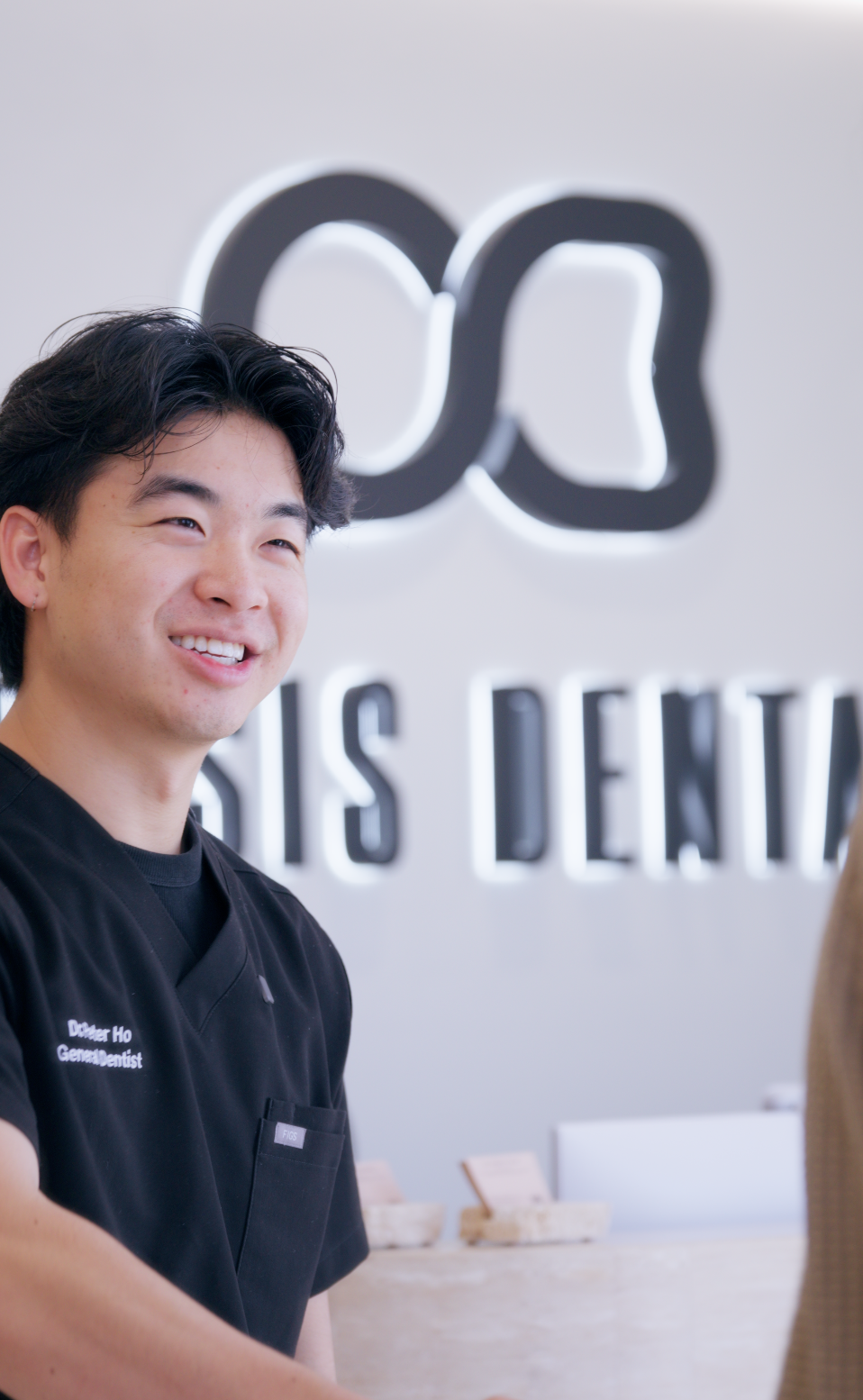 A smiling male dental professional standing inside a dental clinic, with the clinic's logo and the words "DDS Dentistry" displayed in the background.