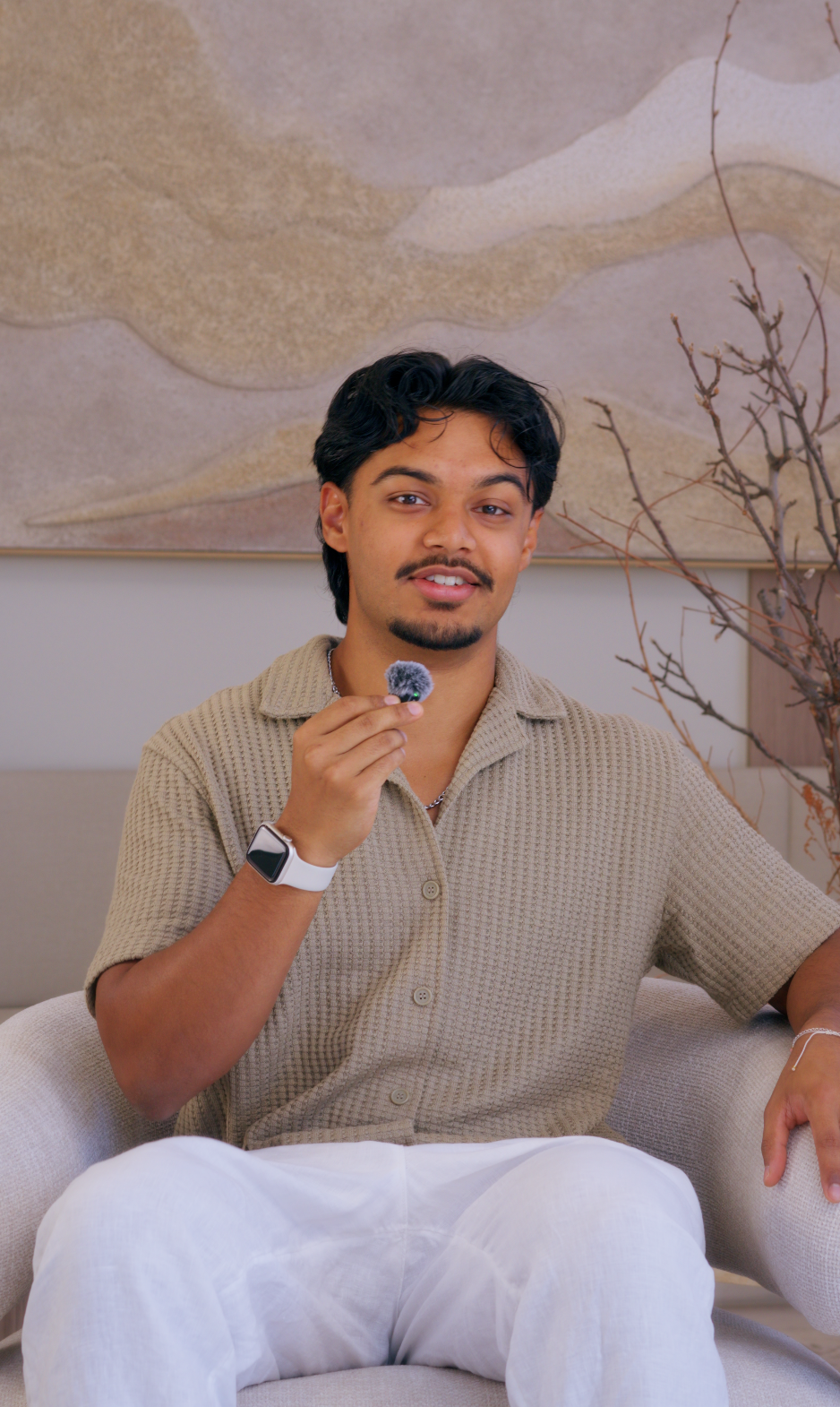 A young man with dark hair and a goatee sitting on a beige couch, holding a mic, wearing a tan short-sleeve shirt and white pants, with a smartwatch on his left wrist, in a cozy room with a large abstract wall art and bare branches in the background.