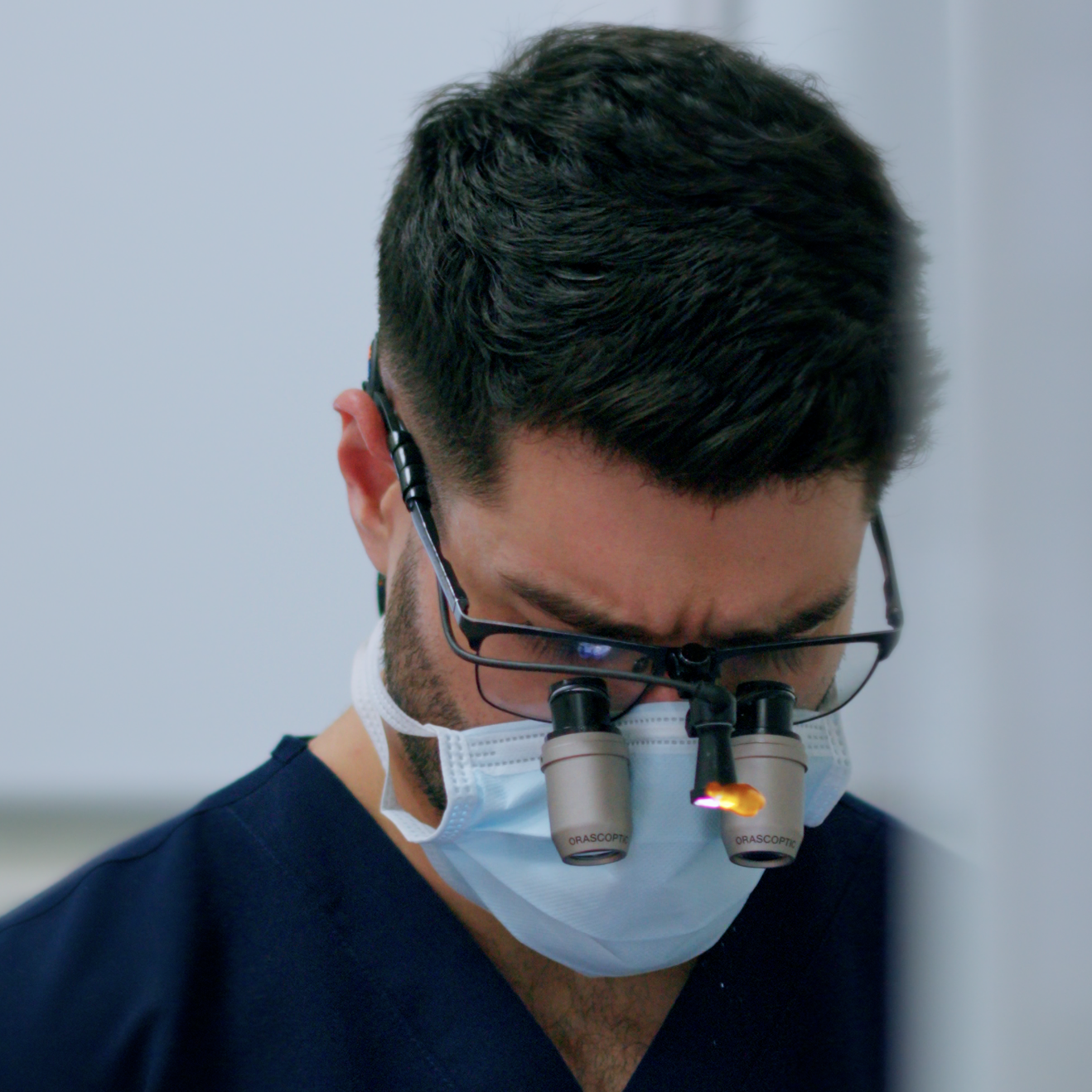 A male healthcare professional wearing magnifying glasses with attached lights, a protective face mask, and blue scrubs, looking down while performing a medical procedure.