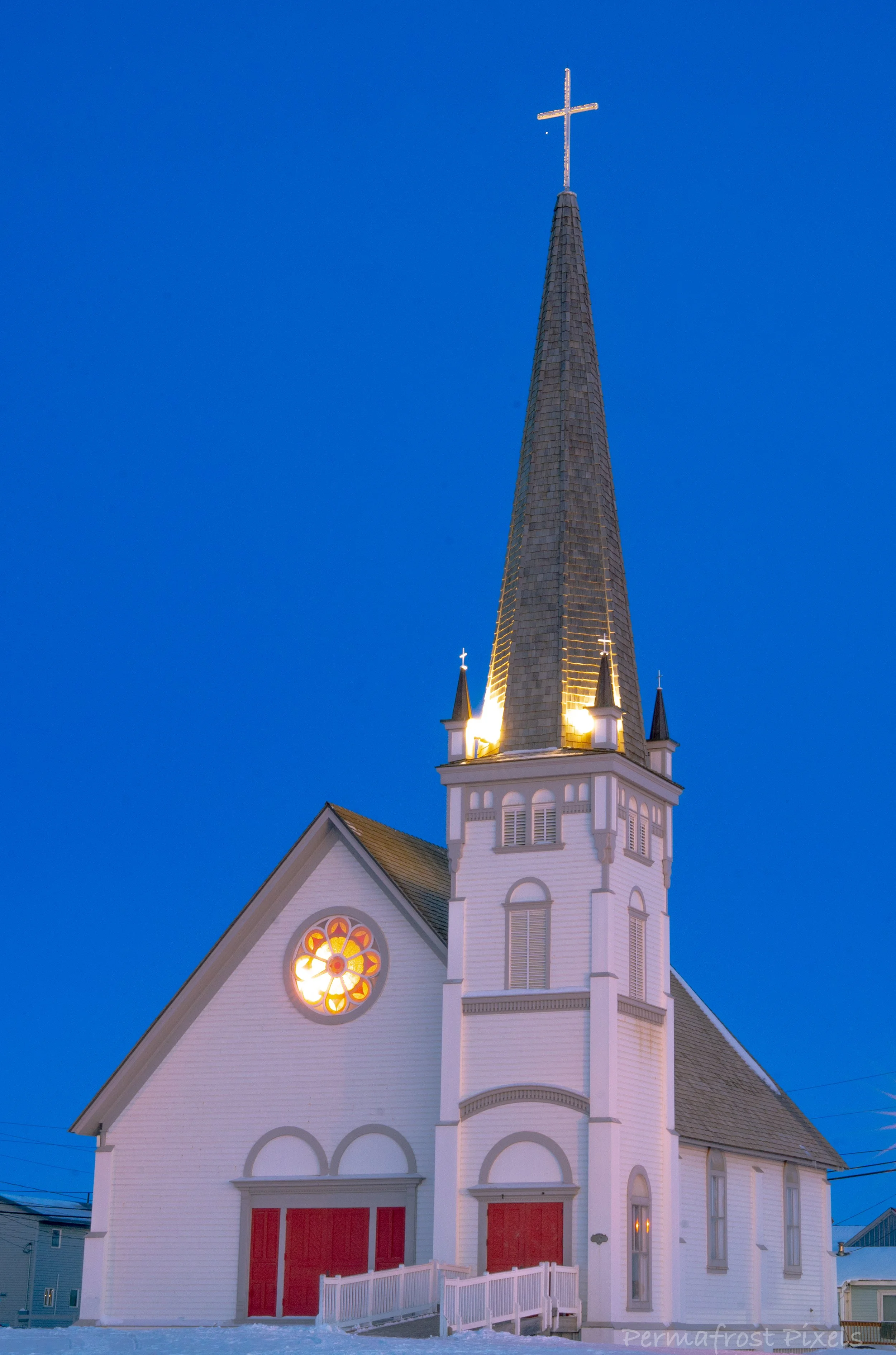 White church with red doors and tall steeple topped with a cross, illuminated against a deep blue evening sky.