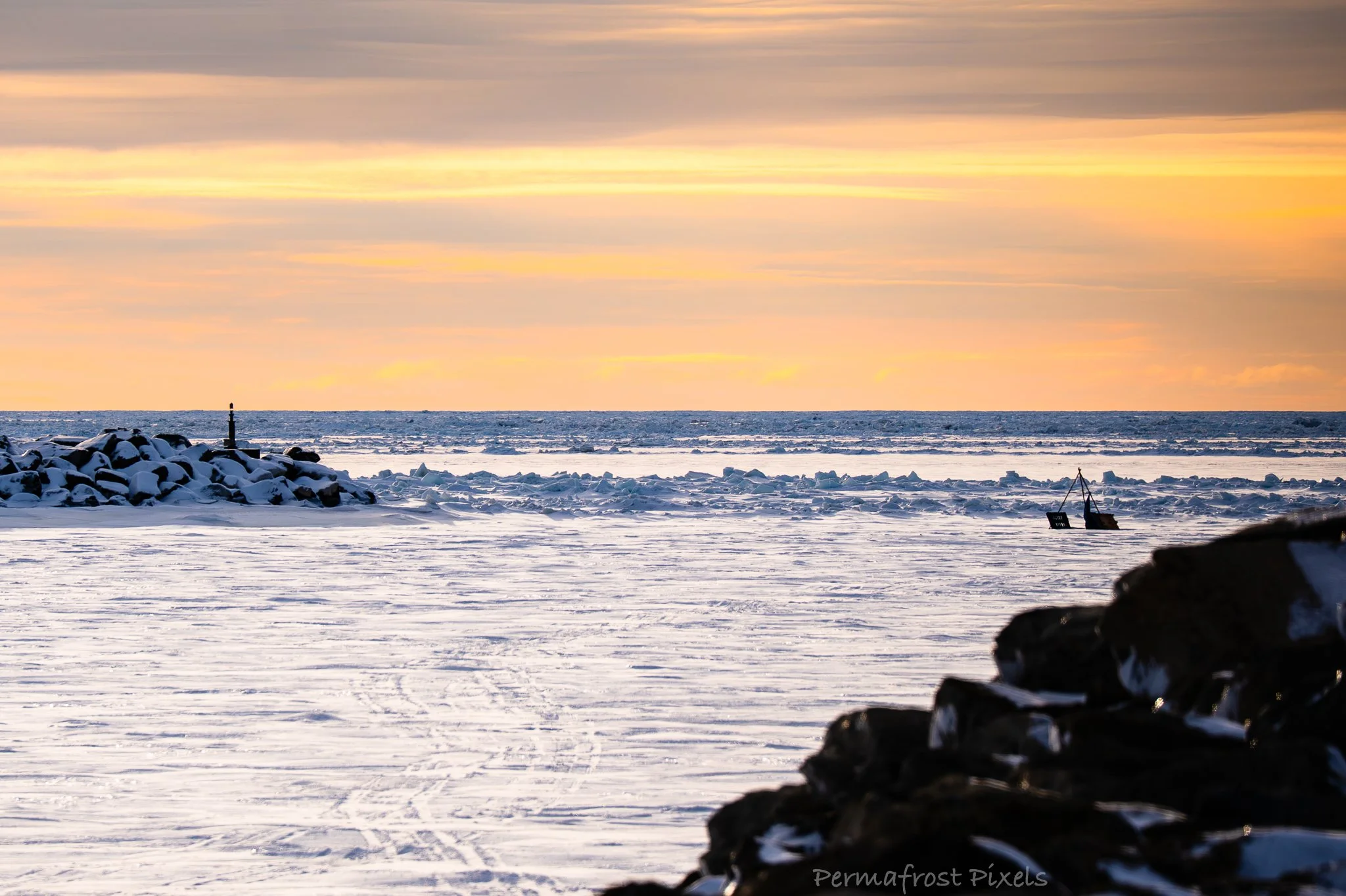 Winter scene at the beach with snow and ice covering the sand, rocks piled on the shoreline, a small lighthouse in the distance, and a colorful sunset sky.