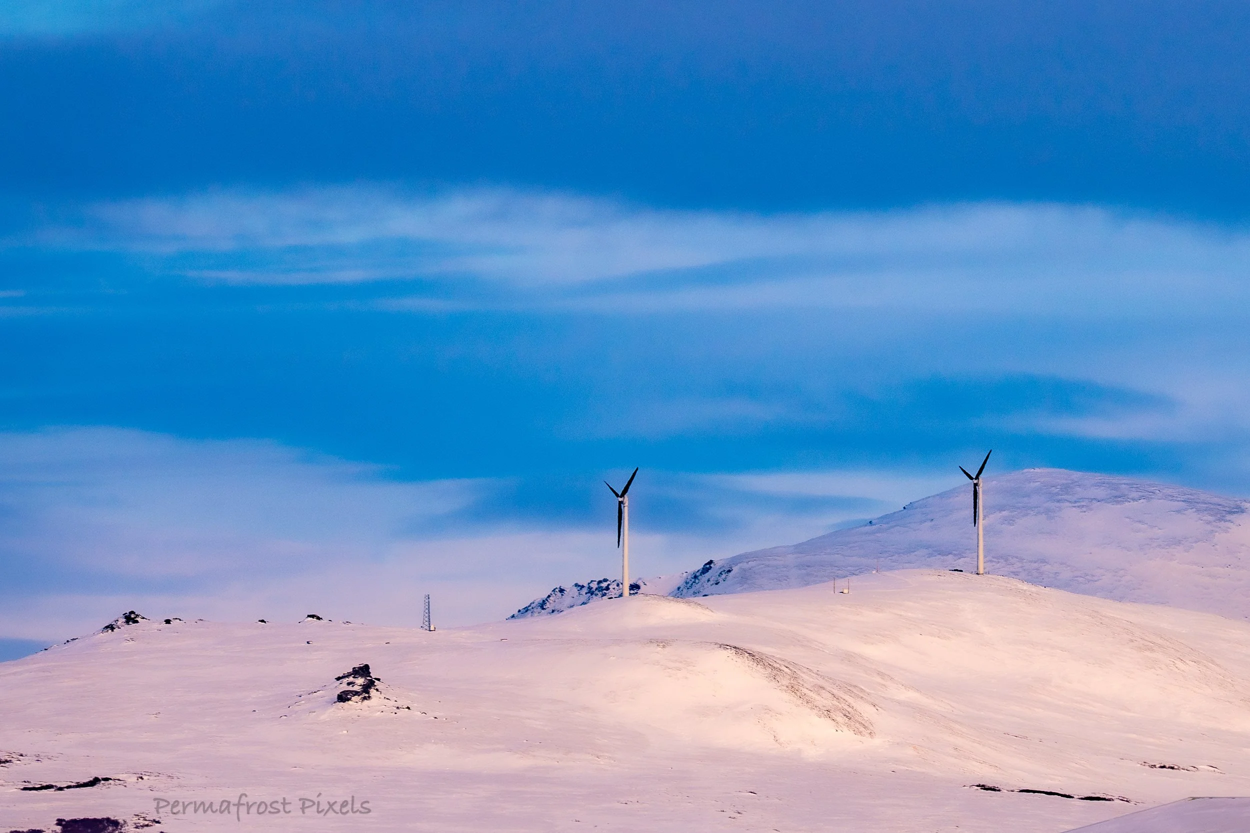 Snow-covered landscape with two wind turbines on a hillside under a partly cloudy sky.
