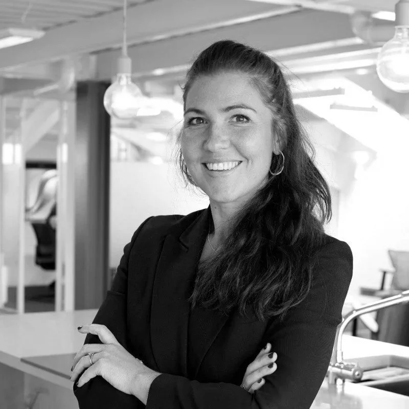 A woman with long dark hair in a ponytail, smiling with arms crossed, dressed in a black blazer, in an office setting with hanging lights and office furniture in the background.