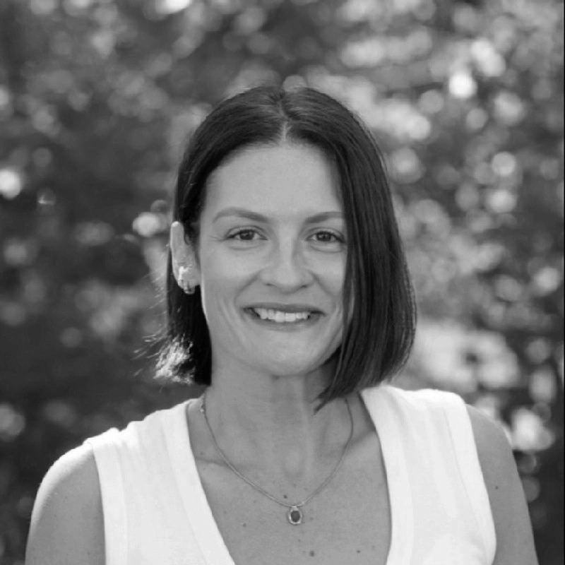 Black and white photo of a woman with short dark hair, smiling, wearing earrings, a necklace with a pendant, and a sleeveless top outdoors.
