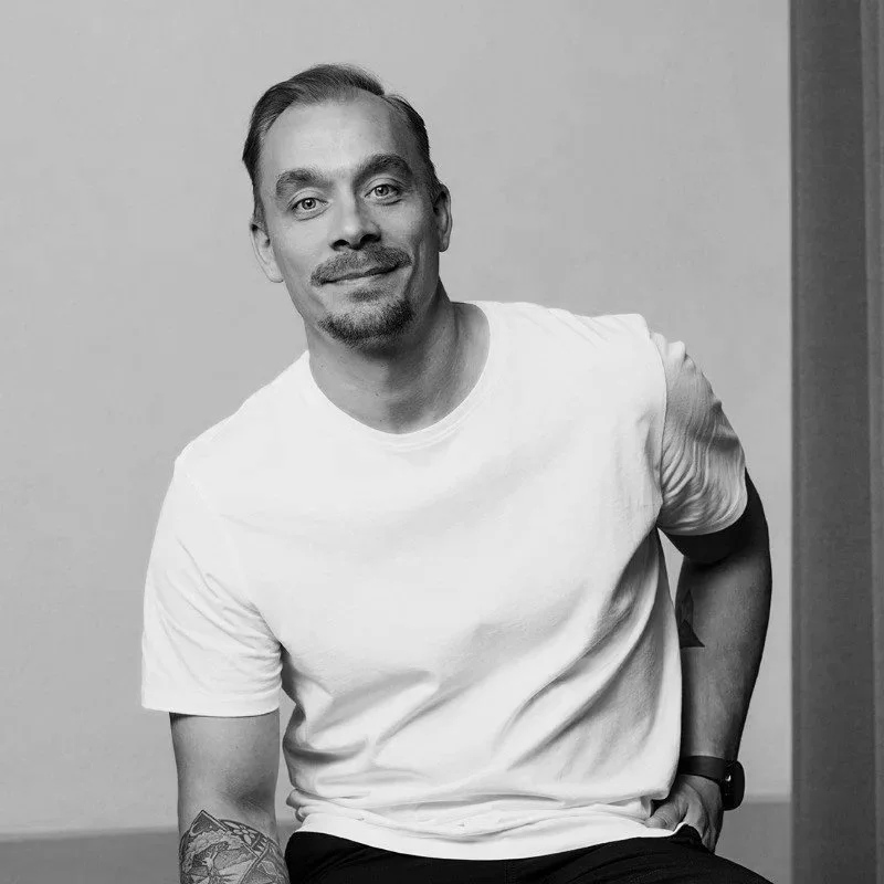 Black and white photo of a smiling man with a goatee and short hair, wearing a white t-shirt and a watch on his left wrist, standing against a plain wall.