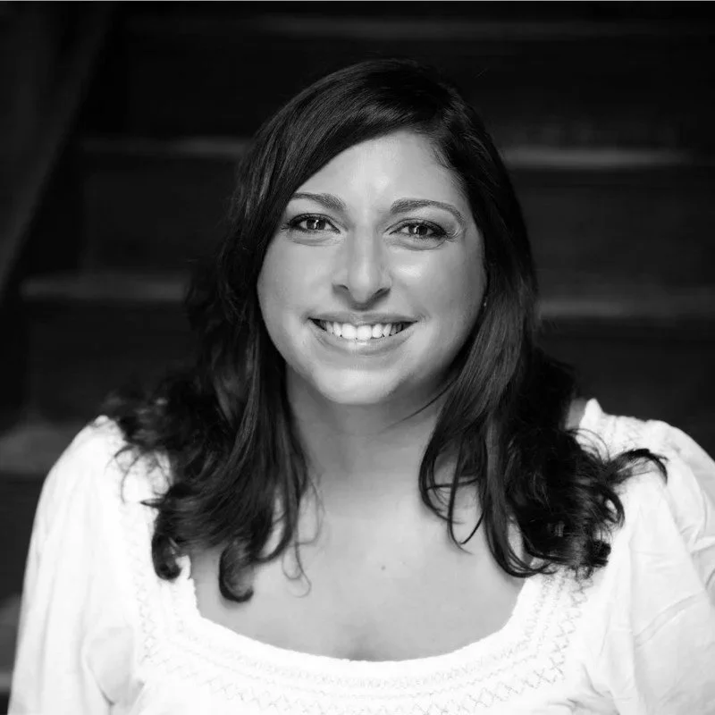 Black and white portrait of a smiling woman with shoulder-length dark hair, wearing a light-colored top, in front of a staircase.
