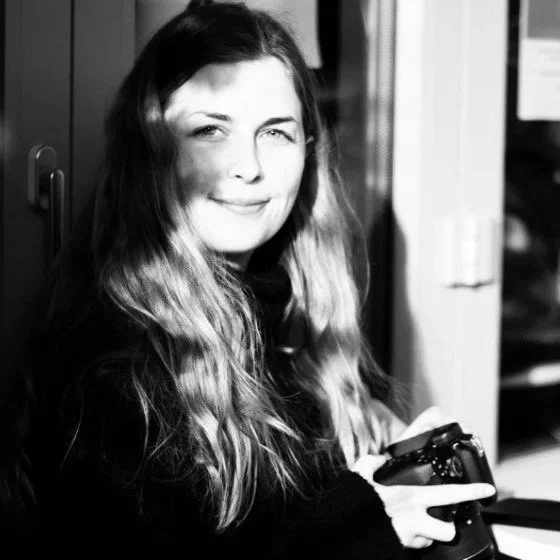A young woman with long, wavy hair is smiling and holding a camera in her hands. She is sitting indoors, near a closet or wardrobe.