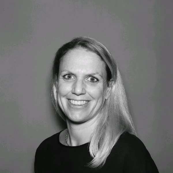 A woman with shoulder-length blonde hair, smiling, wearing a black top, against a plain background.