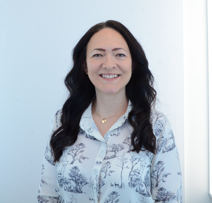 Hayley Wahlbeck with long dark hair smiling, wearing a white blouse with a blue landscape pattern, standing in front of a light-coloured wall.