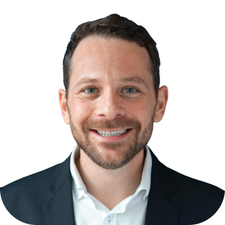 A smiling man with dark hair and a beard, wearing a dark suit jacket and a white collared shirt, against a white background.