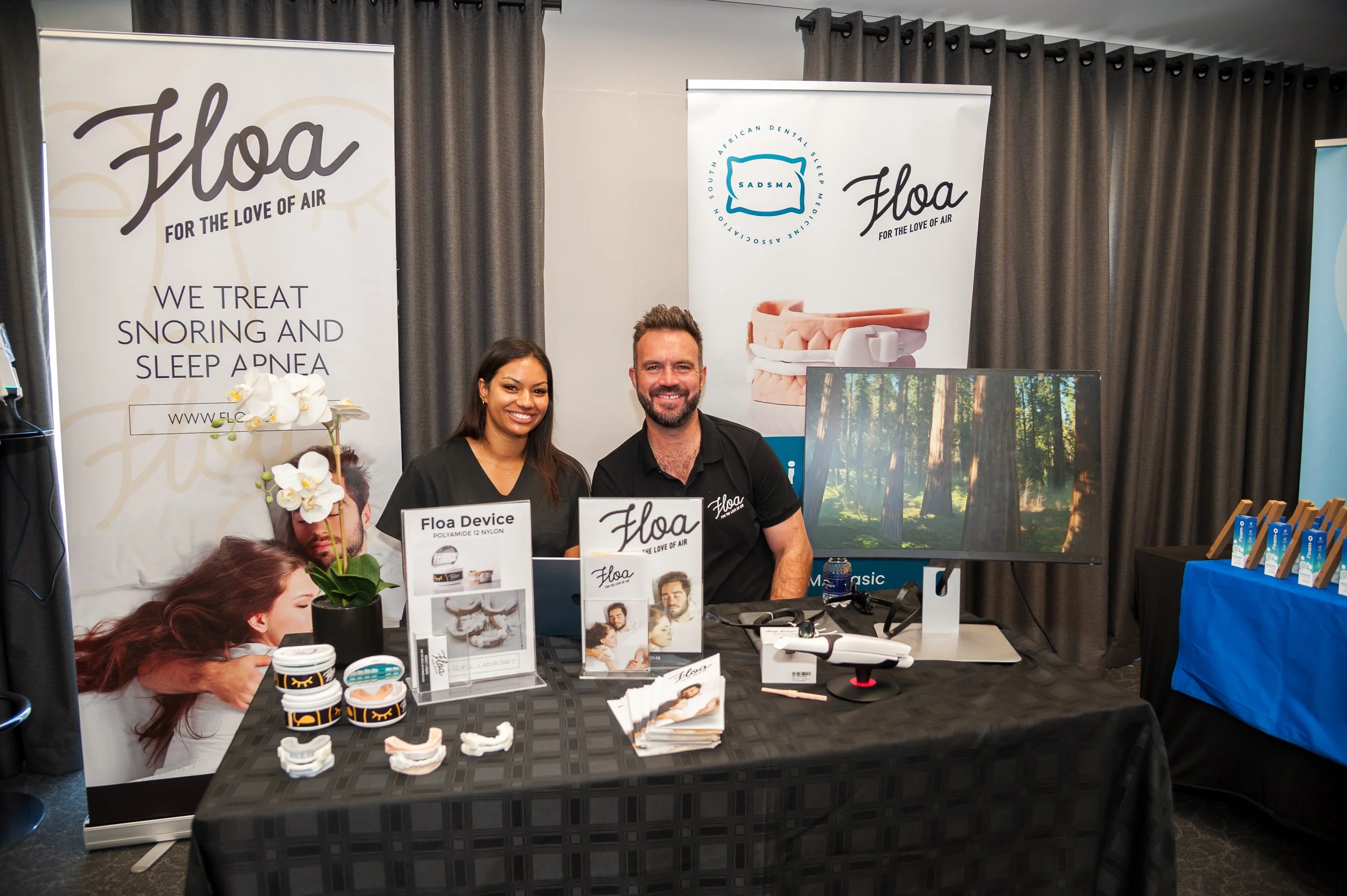 Two people standing behind a display table at a promotional booth for Floa, a company that treats snoring and sleep apnea with devices and solutions. The table has brochures, device models, and a drone, with promotional banners and a television screen showing a forest scene in the background.