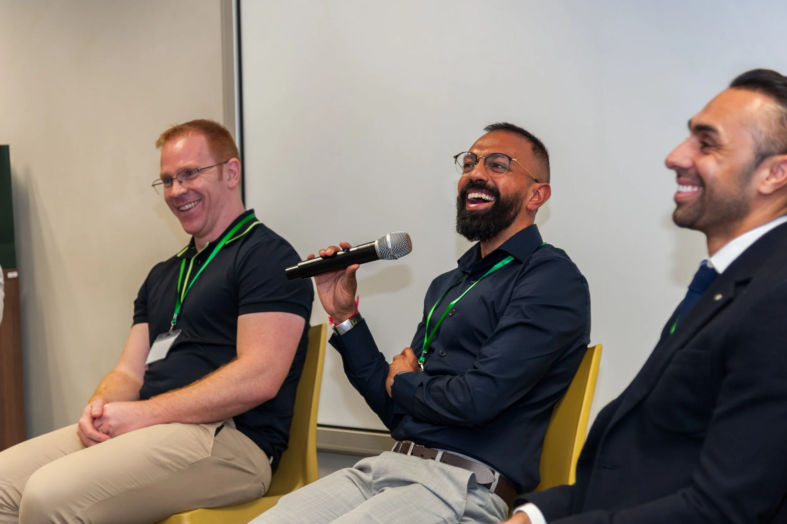 Three men sitting in chairs on a panel, laughing and smiling, with one holding a microphone.