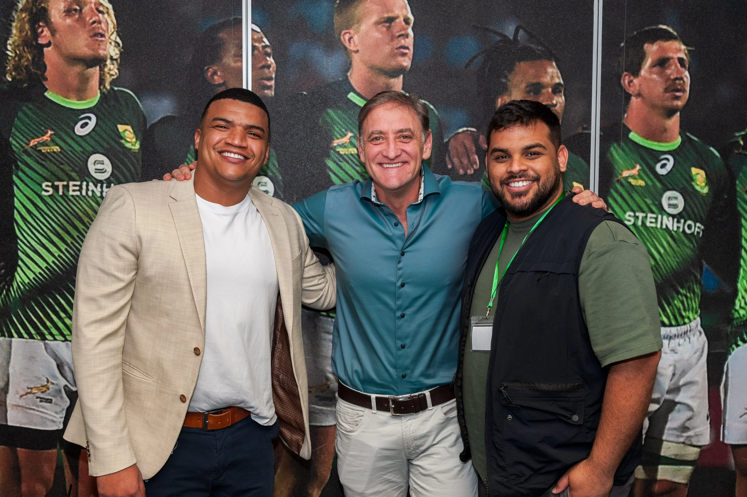 Three men standing together, smiling, with a background featuring rugby players in green and black jerseys.