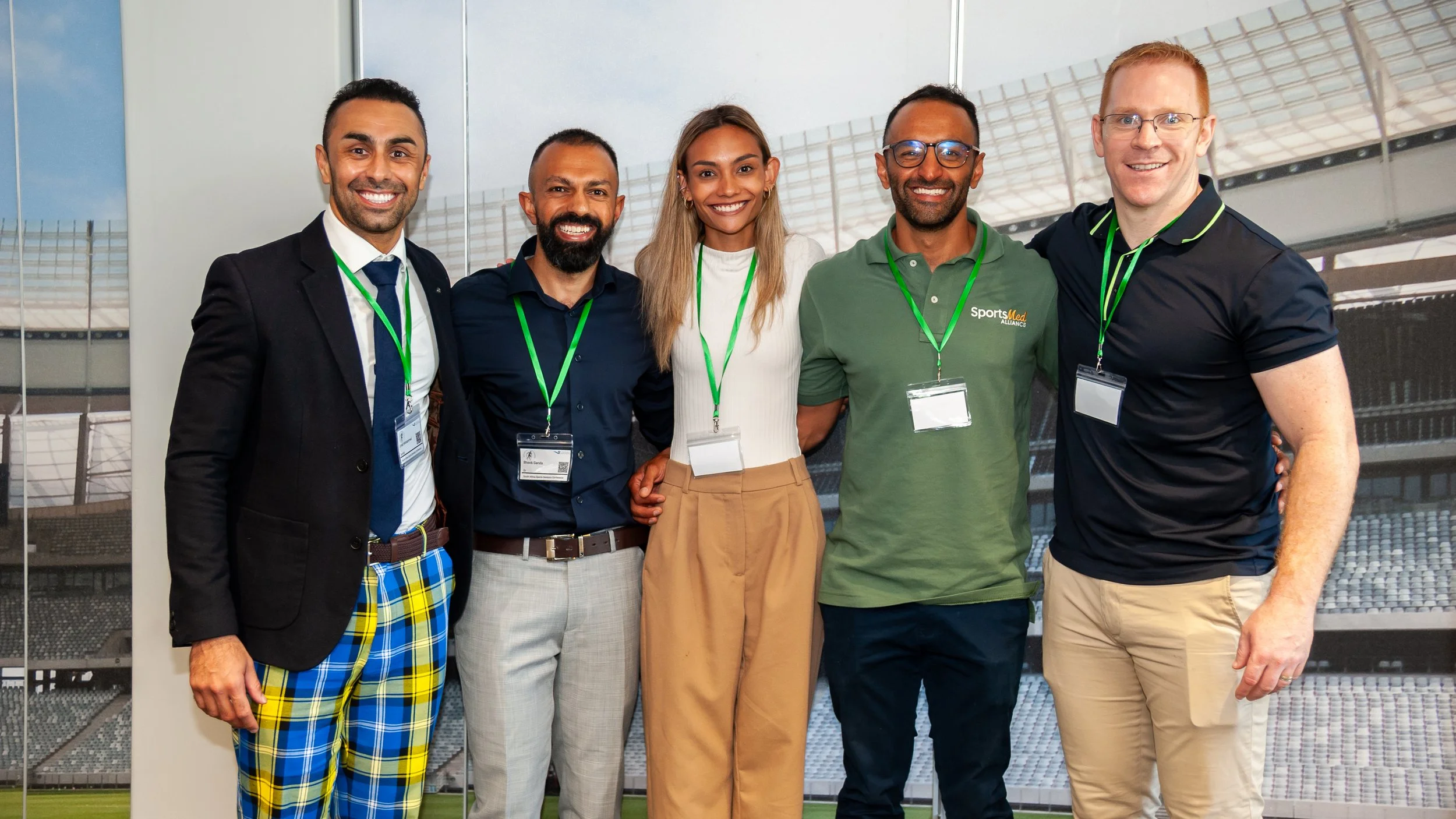 Group of five diverse professionals smiling at the camera, standing inside a stadium with empty seats in the background, all wearing conference badges.