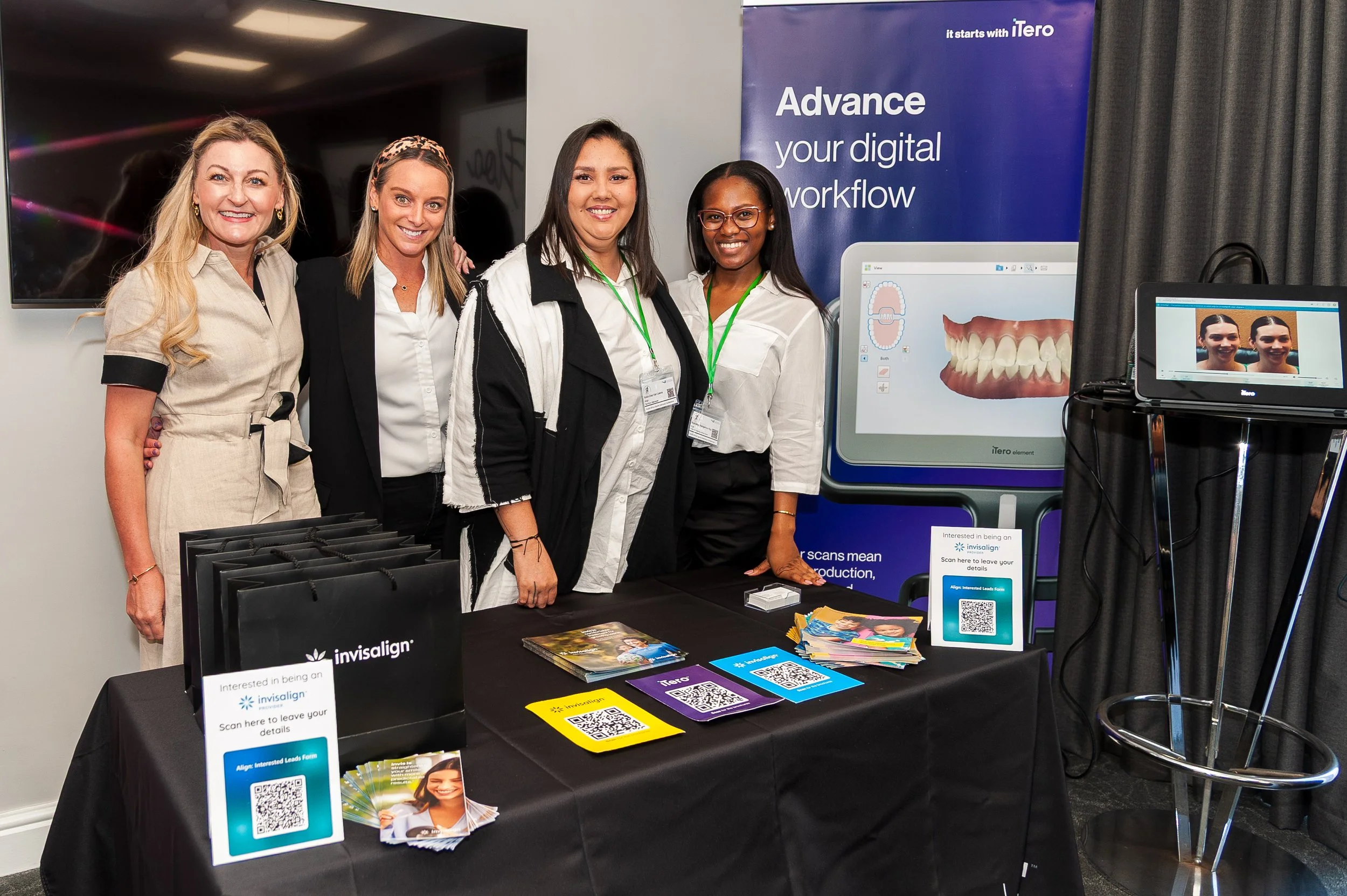 Four women standing behind a promotional table for Invisalign, smiling at the camera, in front of a blue and white sign that reads 'Advance your digital workflow,' with QR codes and pamphlets on the table, and a digital display showing a smile model and red and white teeth.