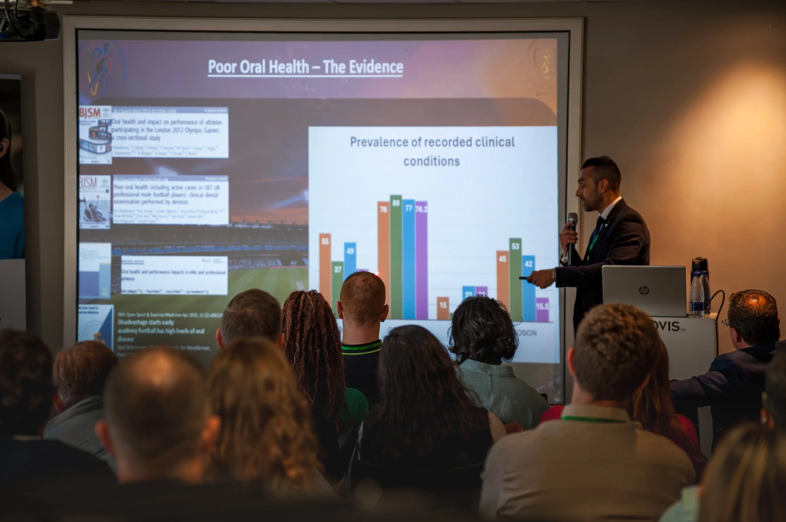 A man in a suit giving a presentation on a projector screen titled 'Poor Oral Health – The Evidence,' with a bar graph showing the prevalence of recorded clinical conditions. Audience members are seated and watching.