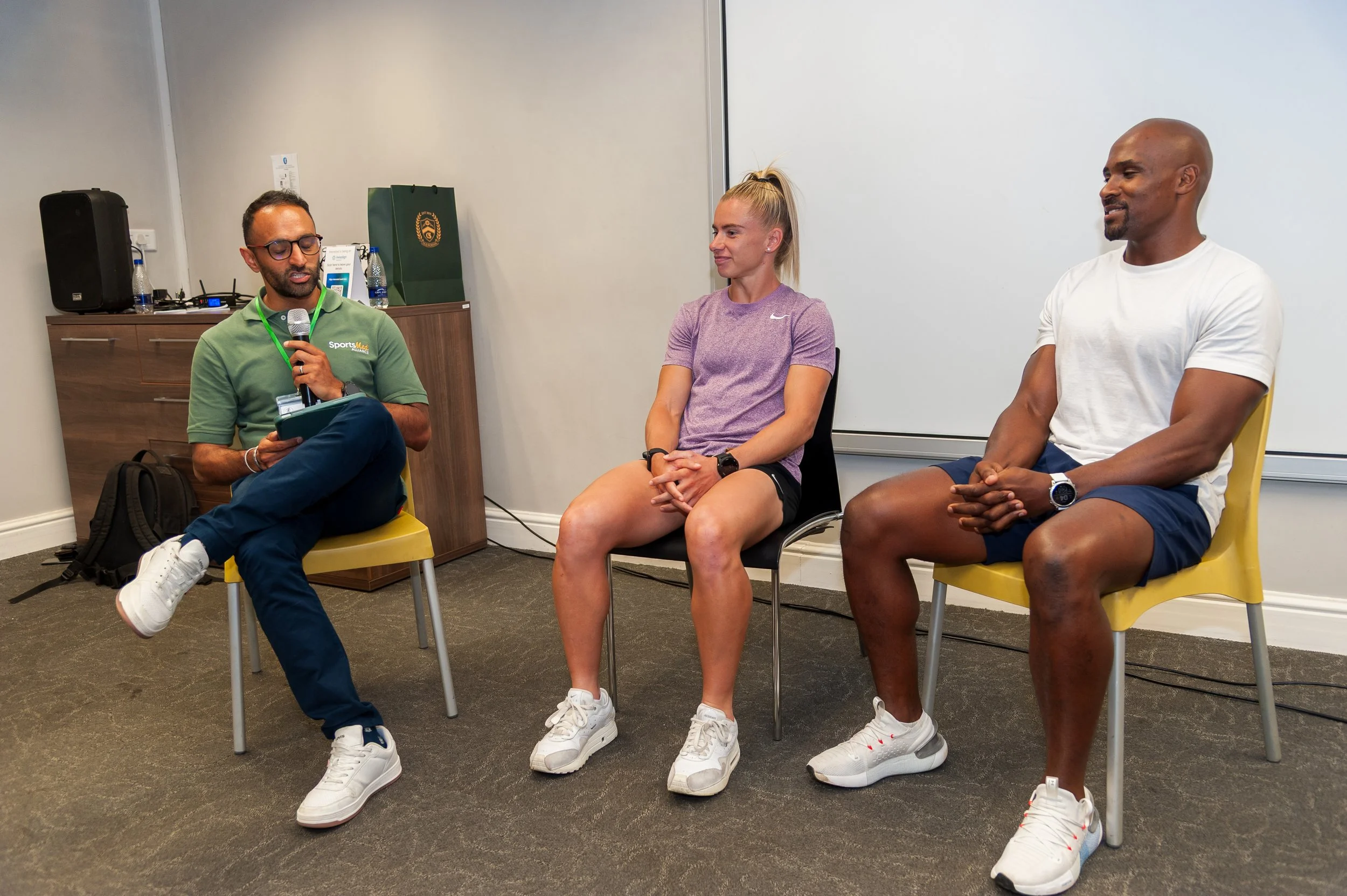 A man in a green polo shirt and glasses is sitting on a yellow chair, holding a microphone and reading from a notepad, during a panel discussion with a woman in a purple athletic shirt and black shorts, and a man in a white t-shirt and navy shorts, both seated on yellow chairs.