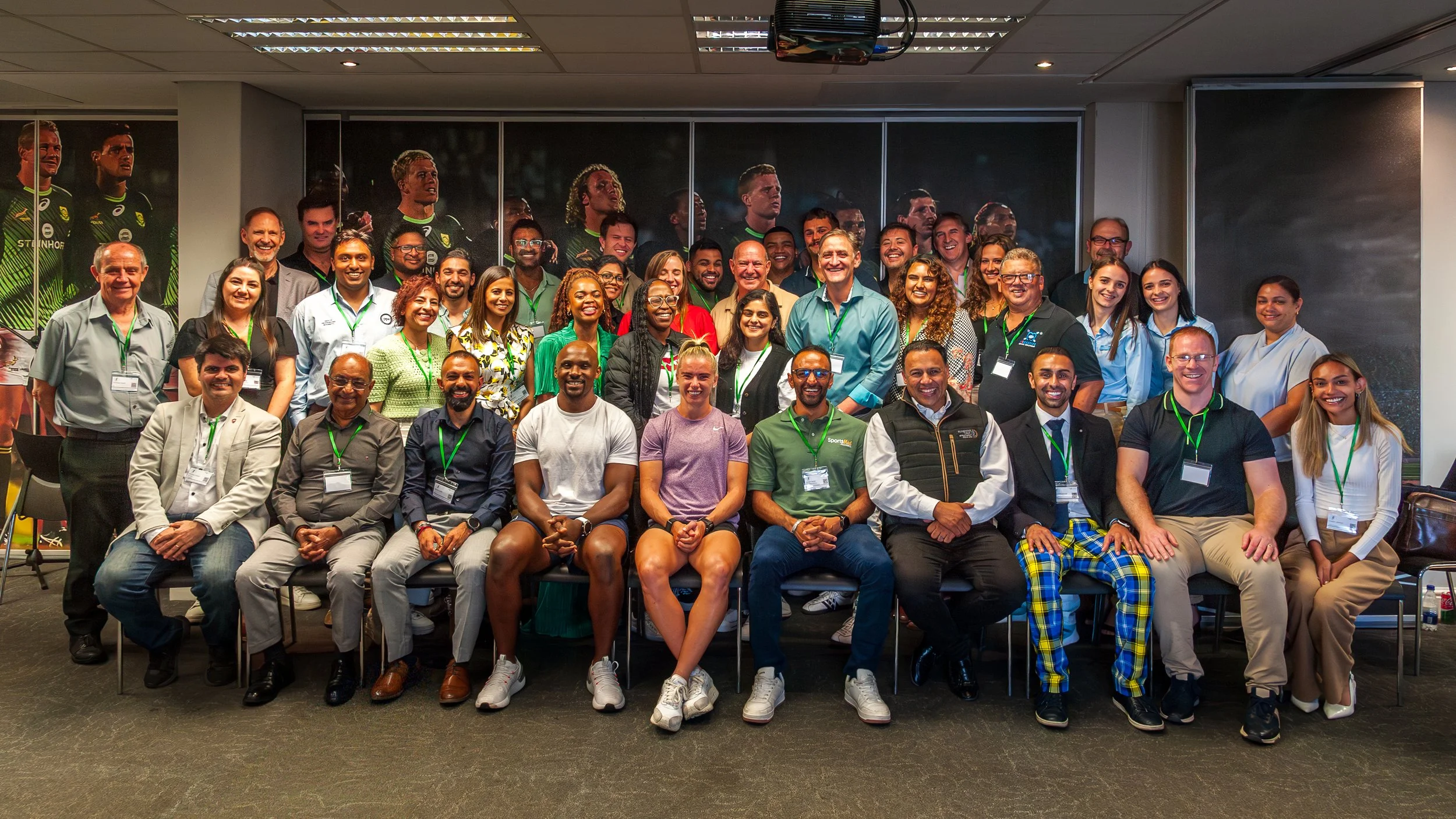 Group of diverse people posing for a photo in an indoor setting, some wearing conference badges, with a backdrop of large sports-themed posters.