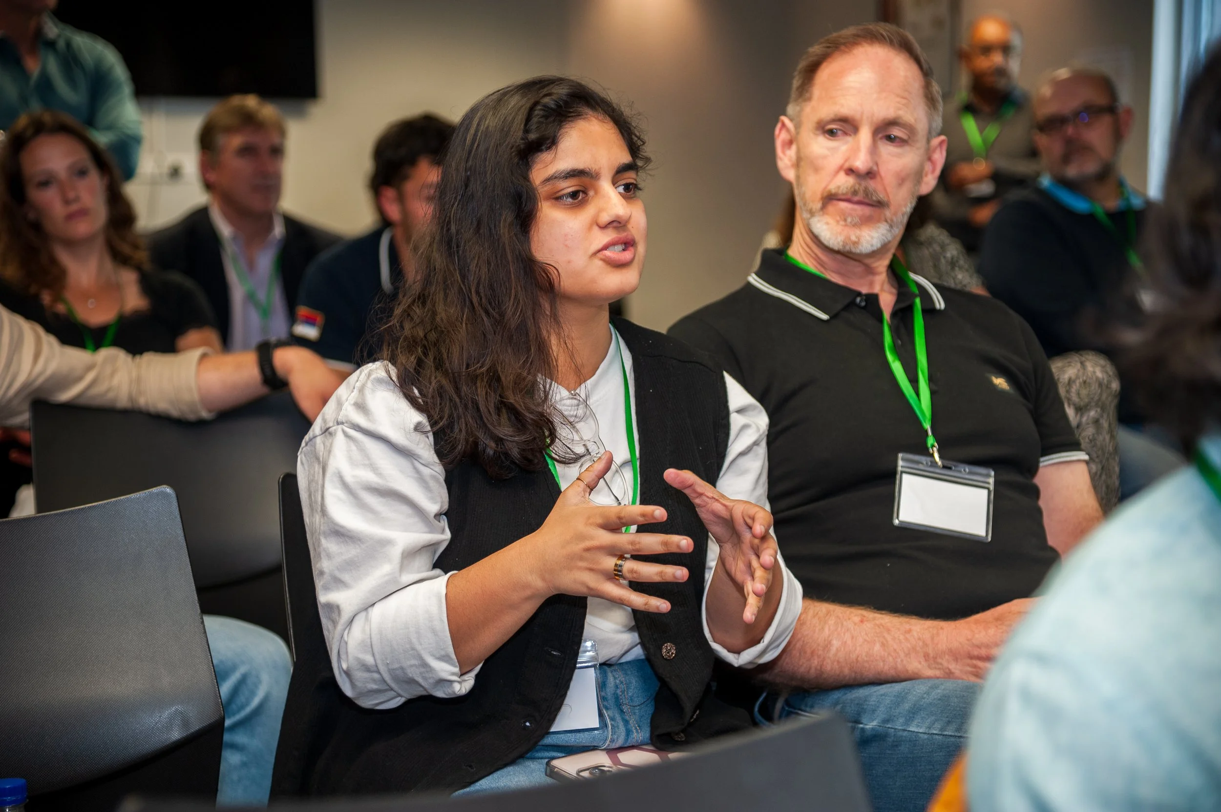 Young woman with long dark hair, wearing a black vest, white shirt, and blue jeans, speaking and gesturing with her hands during a meeting or conference, seated next to an older man with short hair and a beard, wearing a black polo shirt with white trim, both wearing green lanyards with name tags, in a room with other attendees.