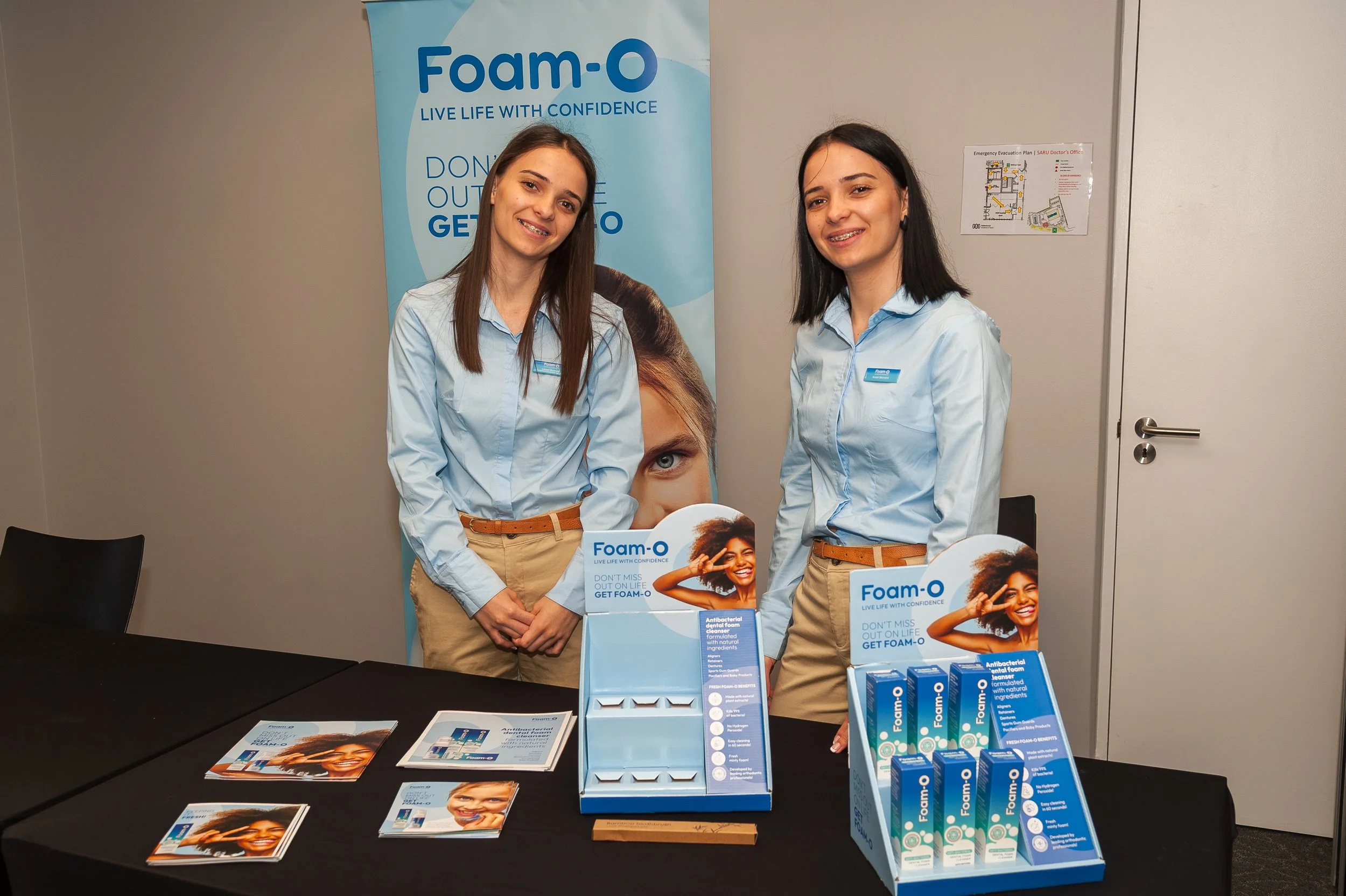 Two women in light blue shirts standing behind a table with Foam-O dental products and promotional materials, with a Foam-O banner in the background.