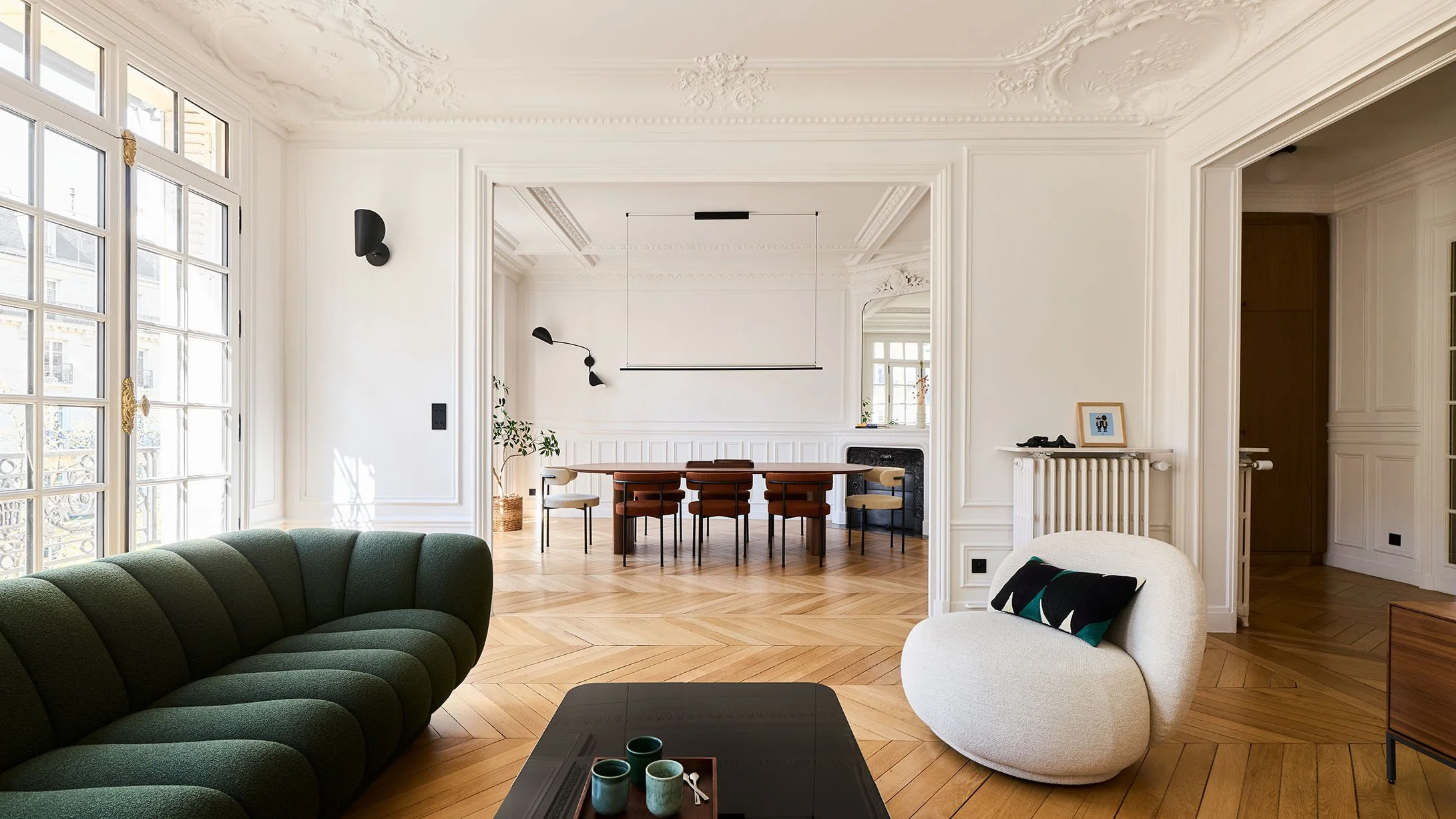 Elegant living room with white walls, ornate ceiling, large windows, green sofa, cream chair with black pillow, black coffee table with three cups, adjacent dining area with wooden table and chairs, and decorative elements.