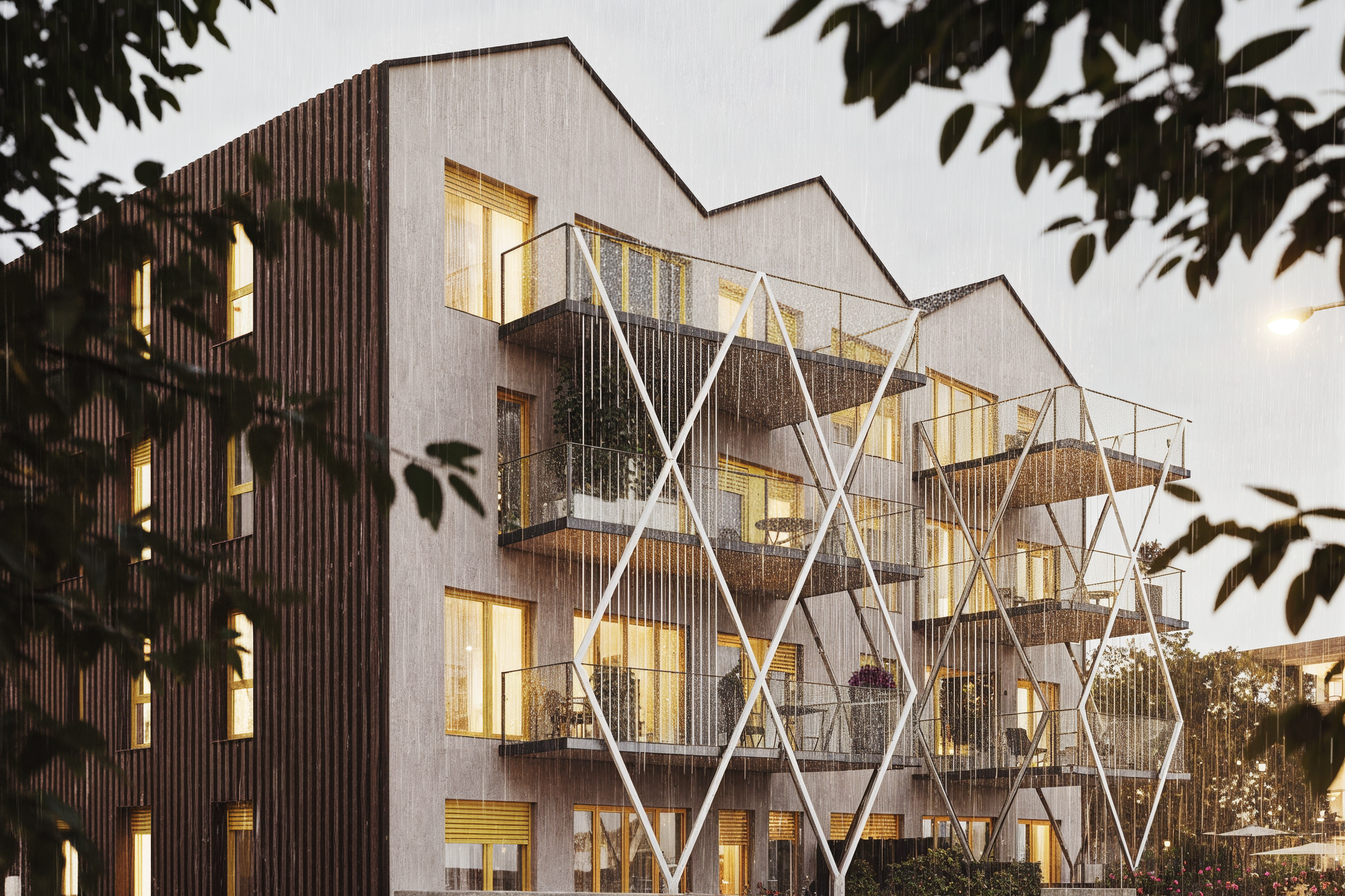 Edificio moderno de varios pisos con balcones y estructura metálica decorativa en la fachada, durante una noche lluviosa con árboles en primer plano.