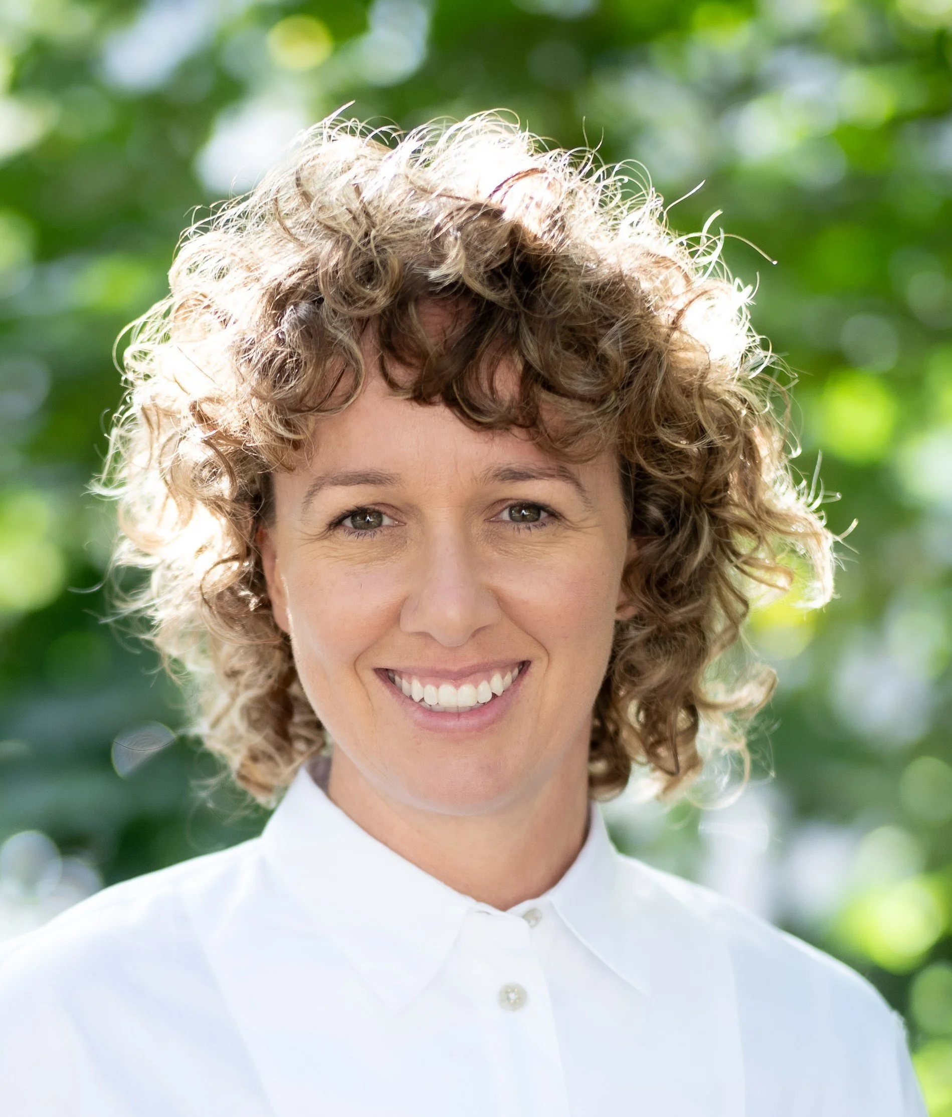 Rebecca Lewis. A woman with curly blonde hair smiling outdoors in front of a green leafy background, wearing a white collared shirt.
