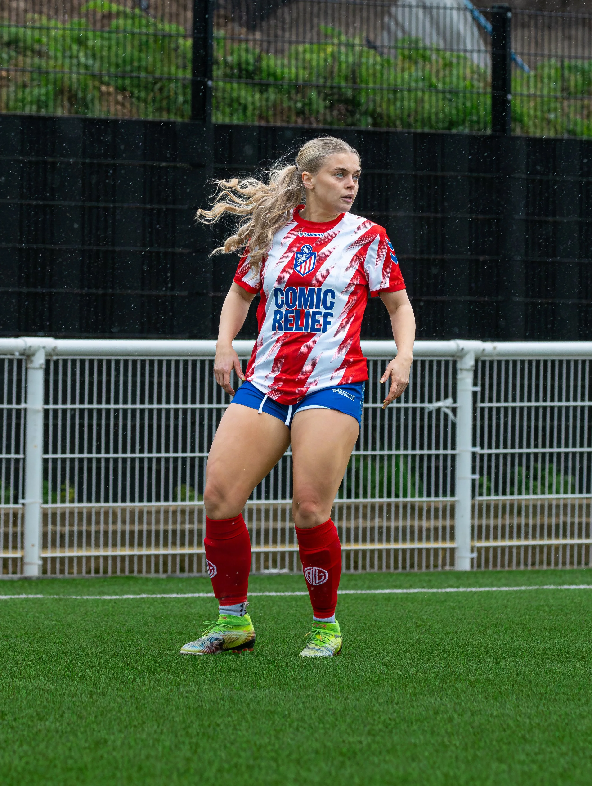 A female soccer player with long blonde hair in a ponytail, wearing a red and white striped jersey with the words 'Comic Relief' and a club logo, blue shorts, red socks, and colorful cleats, is standing on a green soccer field during a rainy day.