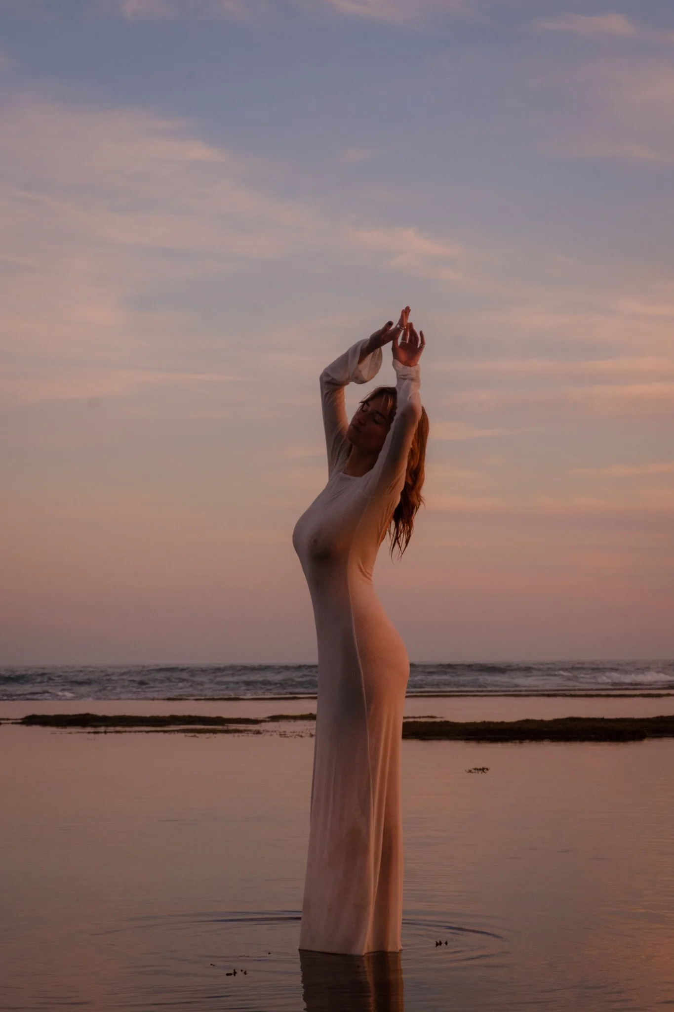 A woman standing in water at sunset with her arms raised above her head, wearing a long, sheer dress.