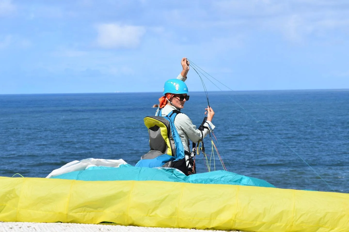 Person wearing a blue helmet, sunglasses, and a life jacket, preparing for paragliding over the ocean.