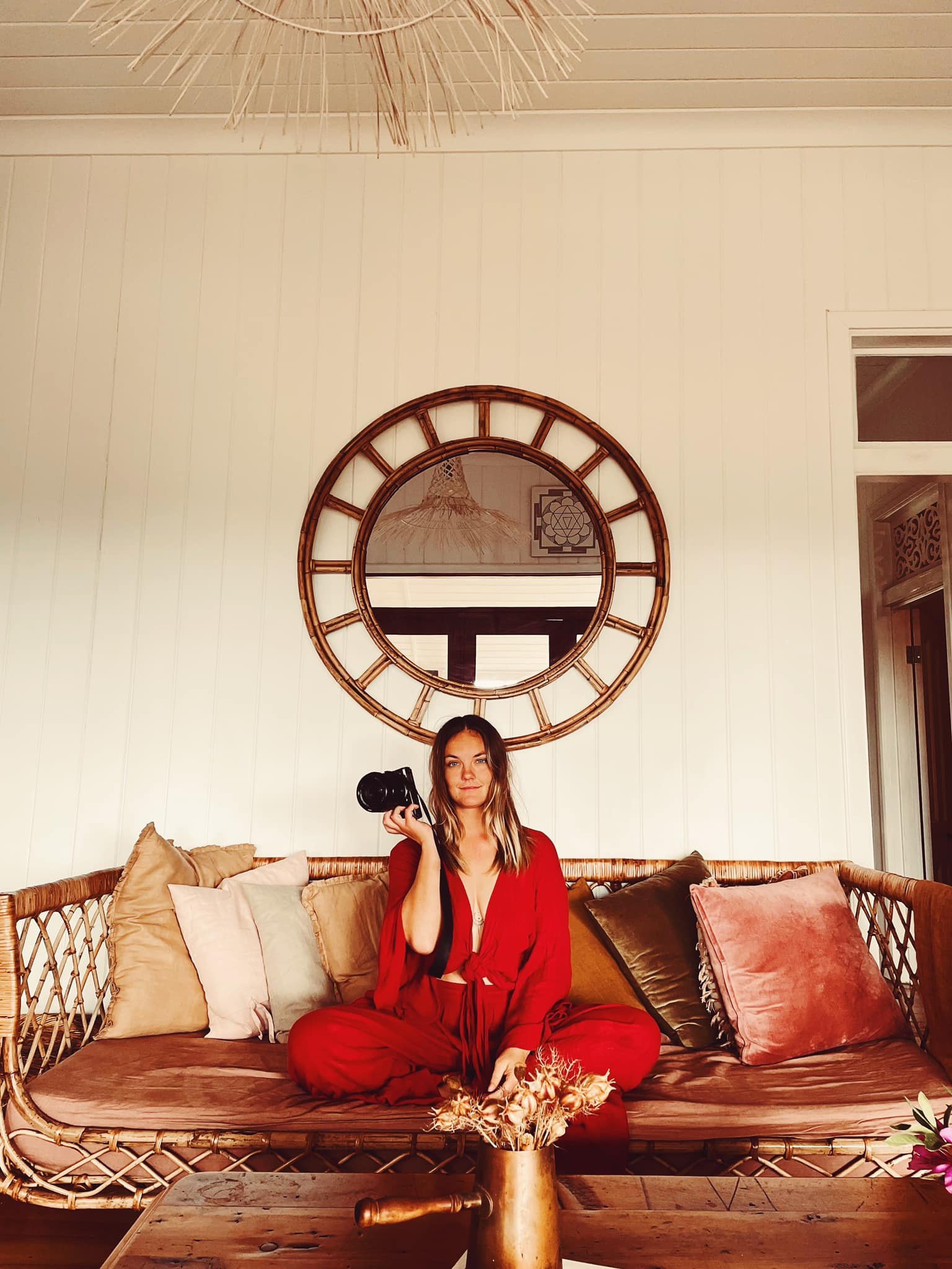 Woman in red outfit sitting on a wicker sofa with pillows, holding a camera, in a room with a large round mirror on the wall and a decorative ceiling light.