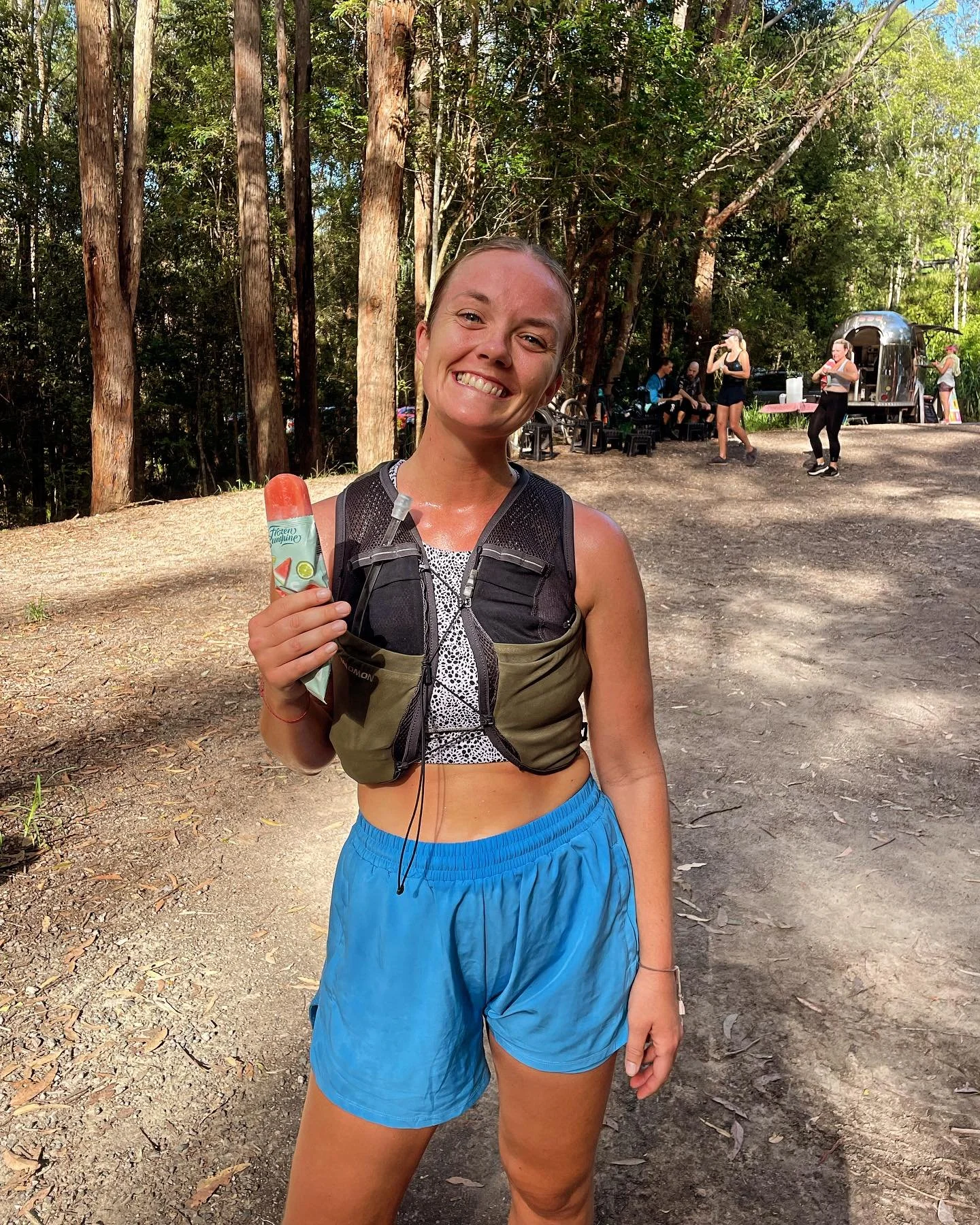 A young woman in athletic clothing smiling outdoors, holding a popsicle, in a wooded area with other people and a trailer in the background.