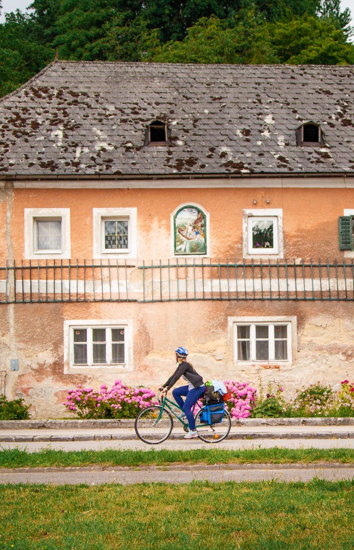 A woman riding a bicycle past an old house with pink flowers in front. The house has four windows, a mural between the windows, and a weathered roof with moss.