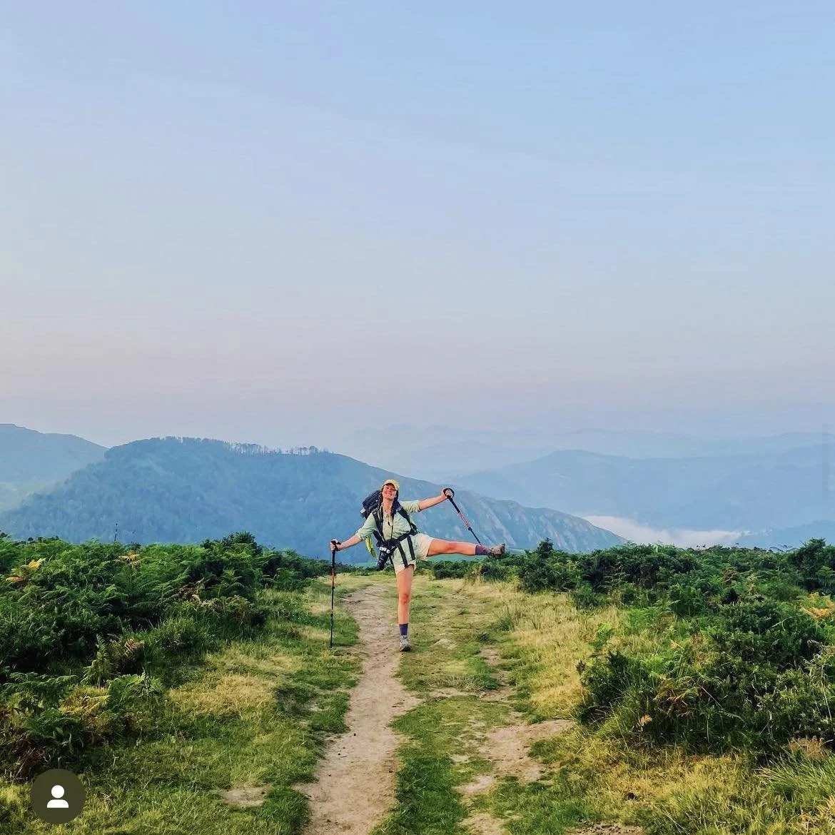 A woman with hiking gear balancing on one leg while holding hiking poles on a trail with mountains and a cloudy sky in the background.