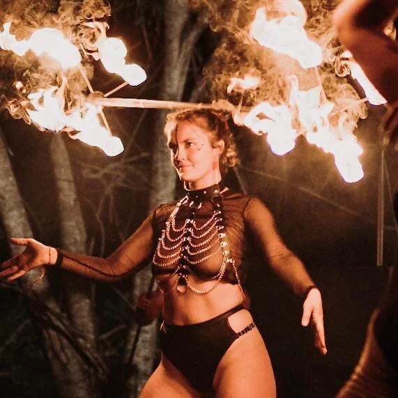 A woman performs a fire breathing act outdoors at night, with flames erupting from her mouth as she suppresses a fire on a stick. She is dressed in black with beaded jewelry and surrounded by a dark environment with trees.