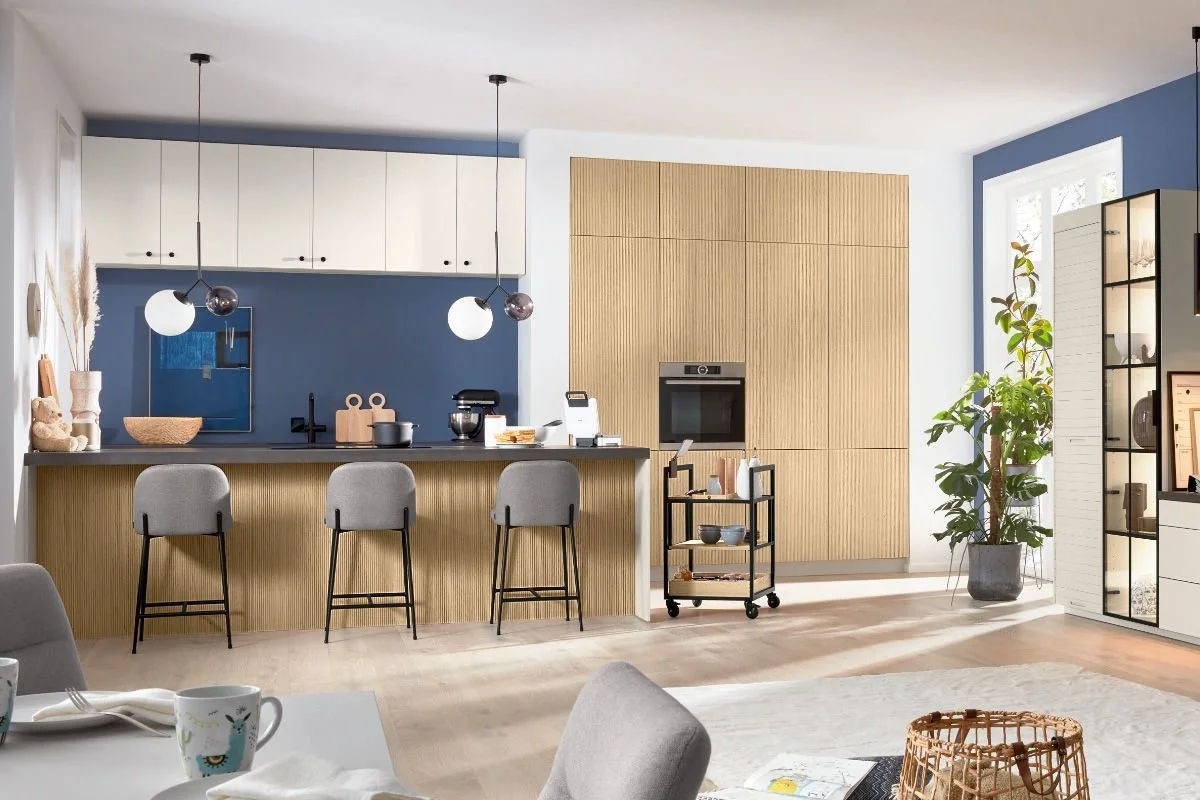 Modern open-concept kitchen and living space with blue and natural wood accents, a kitchen island with three barstools, pendant lighting, and a large plant near windows.