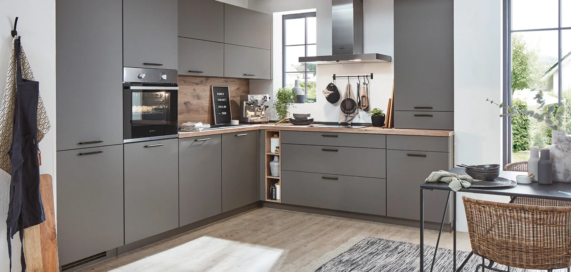 Modern kitchen with gray cabinets, wooden countertops, and large windows. Contains oven, coffee machine, hanging utensils, plants, and a small table with bowls and decorative vases. Sunlight streams in through the windows.