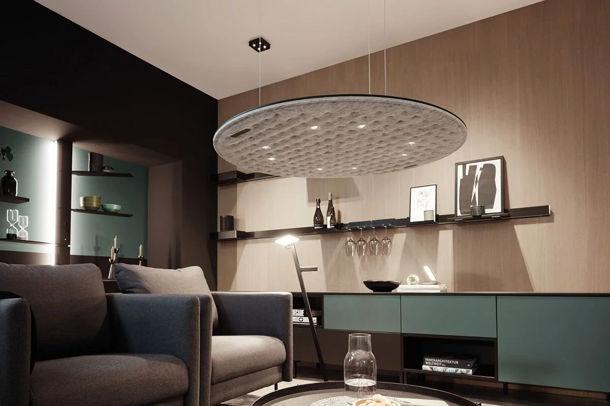 A modern living room with a dark gray sofa, a black and teal cabinet, floating shelves with decorative items, and a circular ceiling speaker or light fixture.